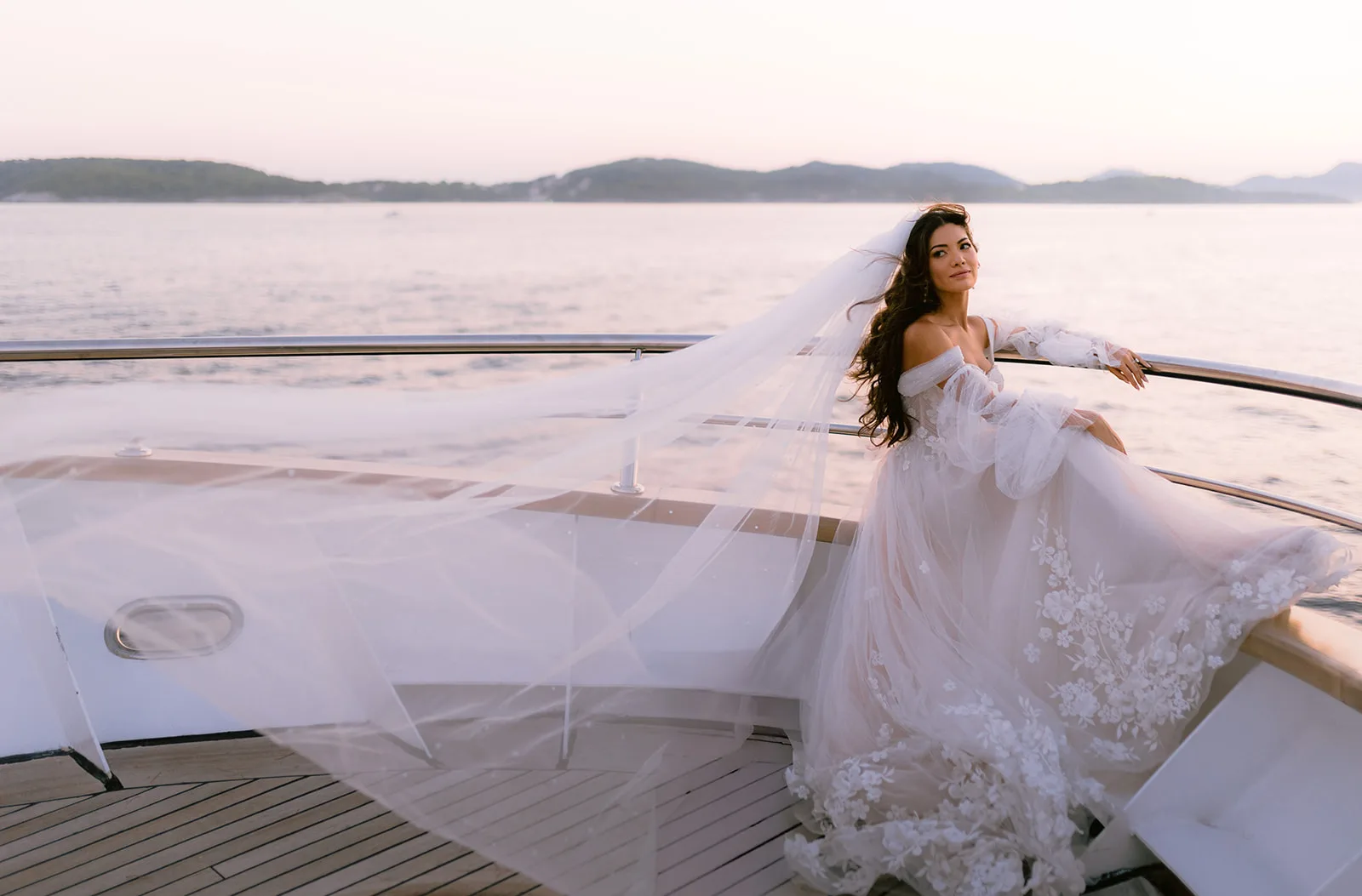 Bride leans against a yacht's chrome railing as her veil billows across the teak deck in golden light.