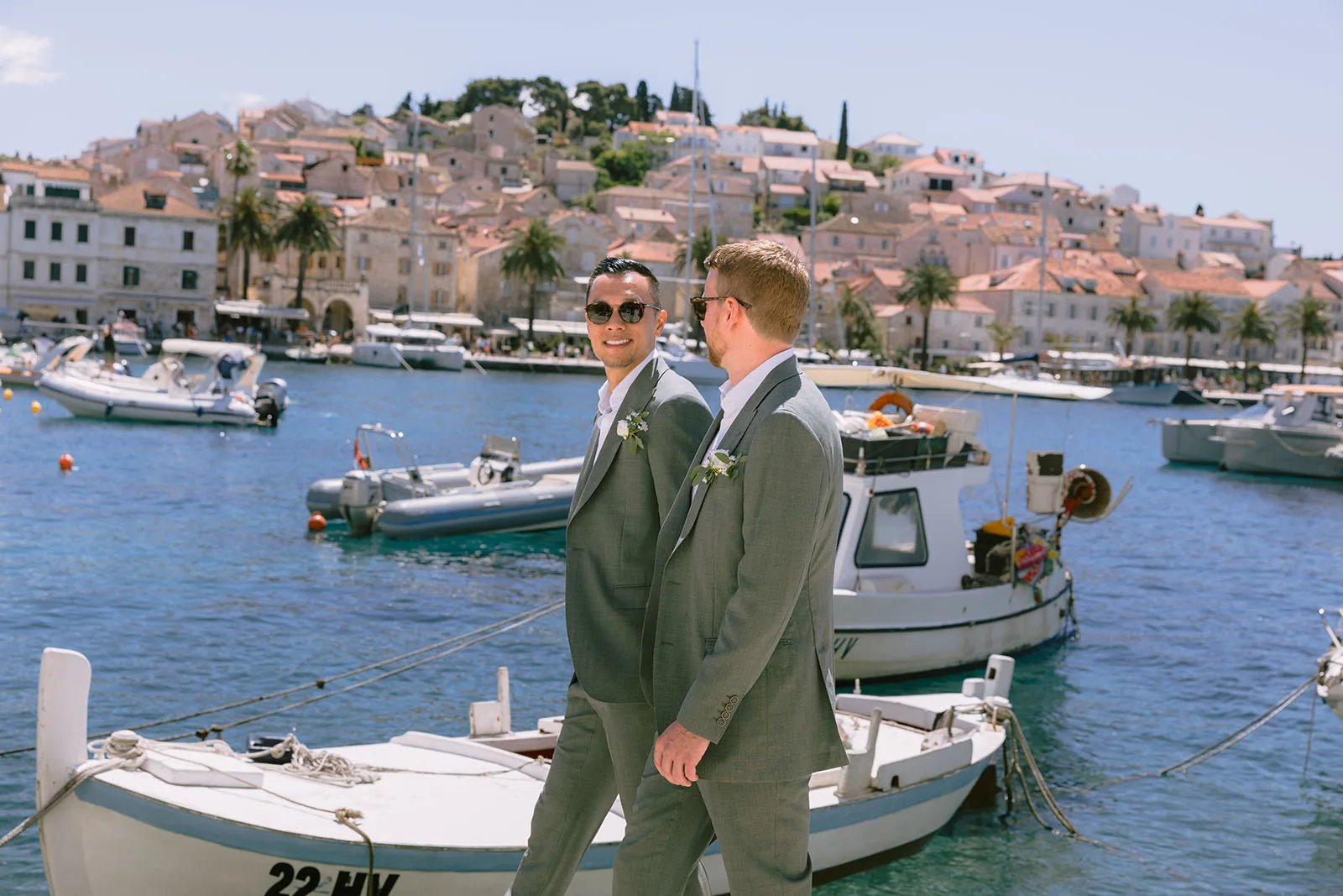 Two grooms in matching grey suits and sunglasses walk along Hvar's stone harbour quay past a moored wooden fishing boat marked "22-HV"; one looks back over his shoulder smiling.