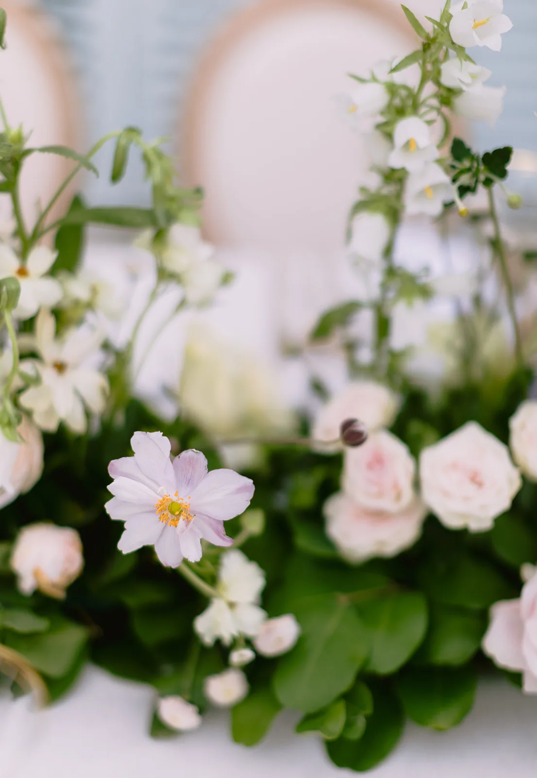 Extreme macro of a floral centerpiece: a pale lavender anemone with a yellow-green center stands out among blush garden roses and sprigs of small white bell-shaped blossoms, framed by greenery.