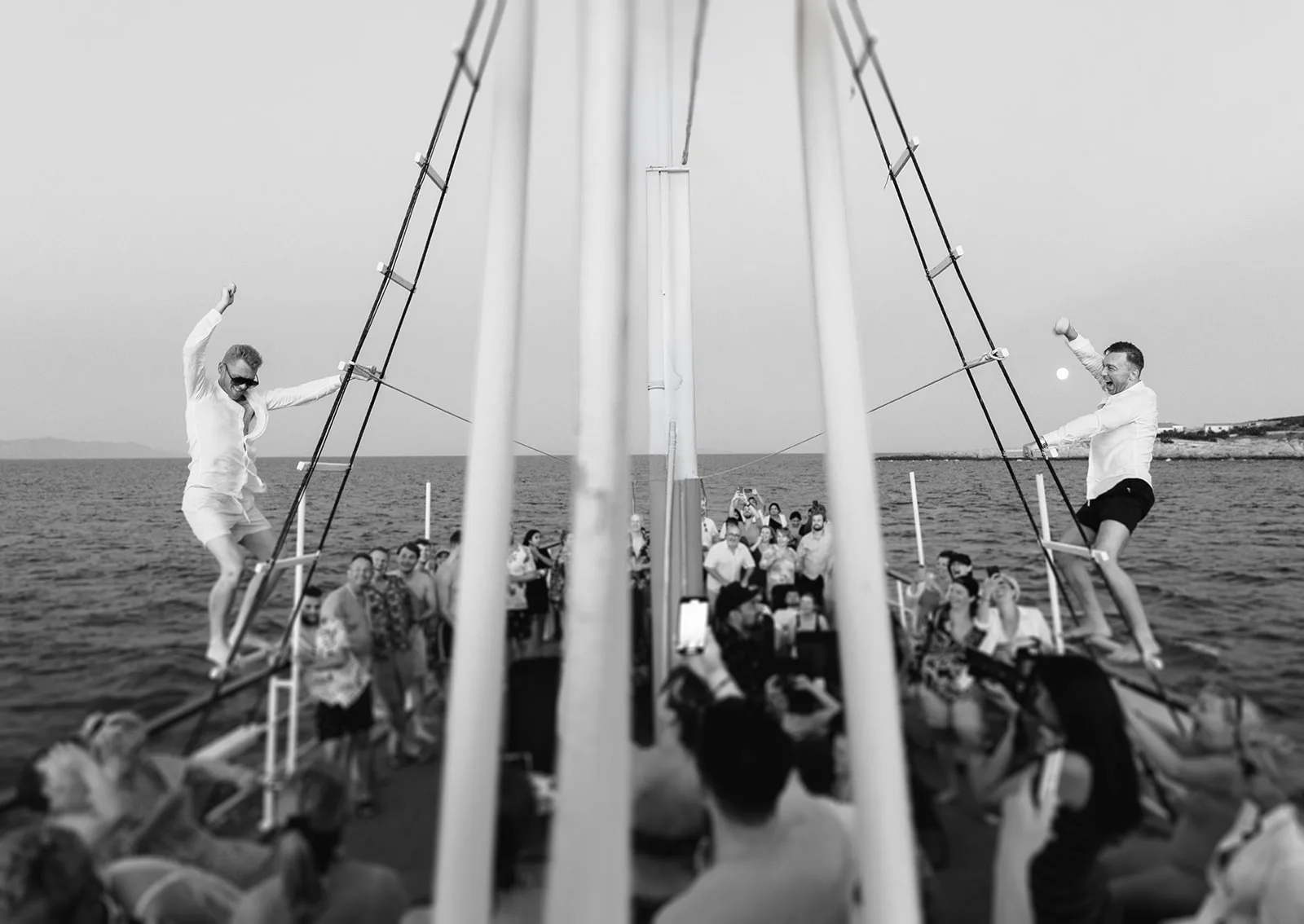 Both grooms scale the boat's rigging in a burst of celebration, arms raised, while a packed crowd of guests cheers below on the sailing deck.