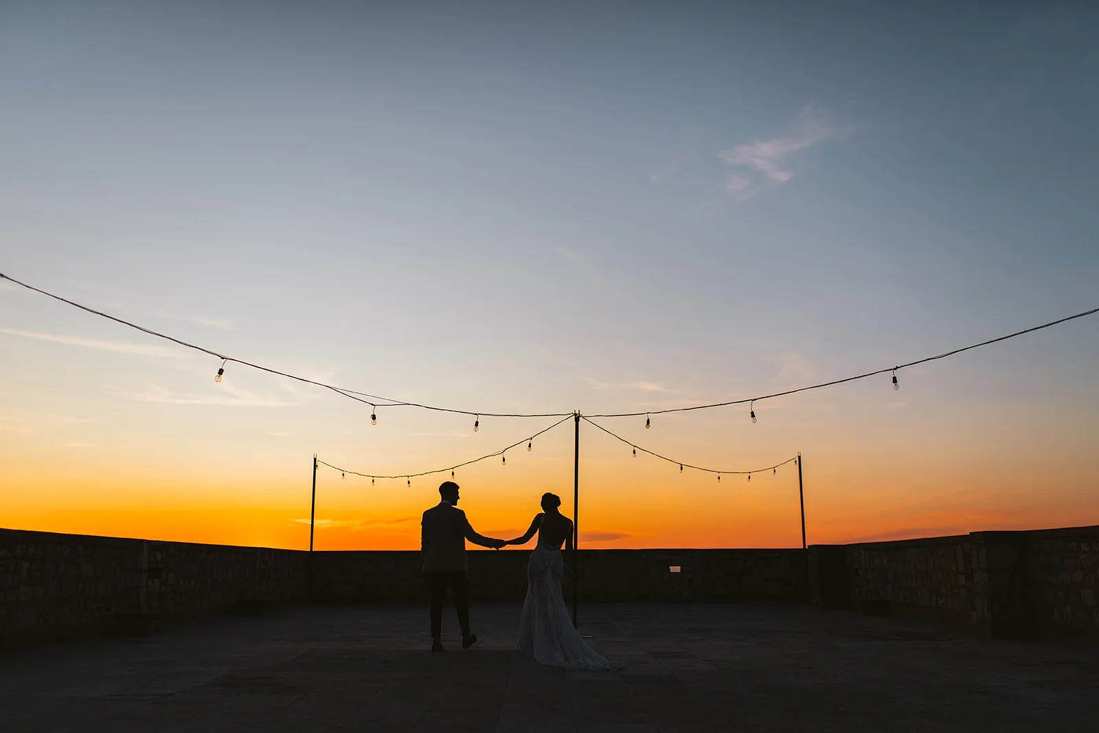 Bride and groom stand in pure silhouette beneath looping string lights as a vivid orange sunset fills the sky behind them.