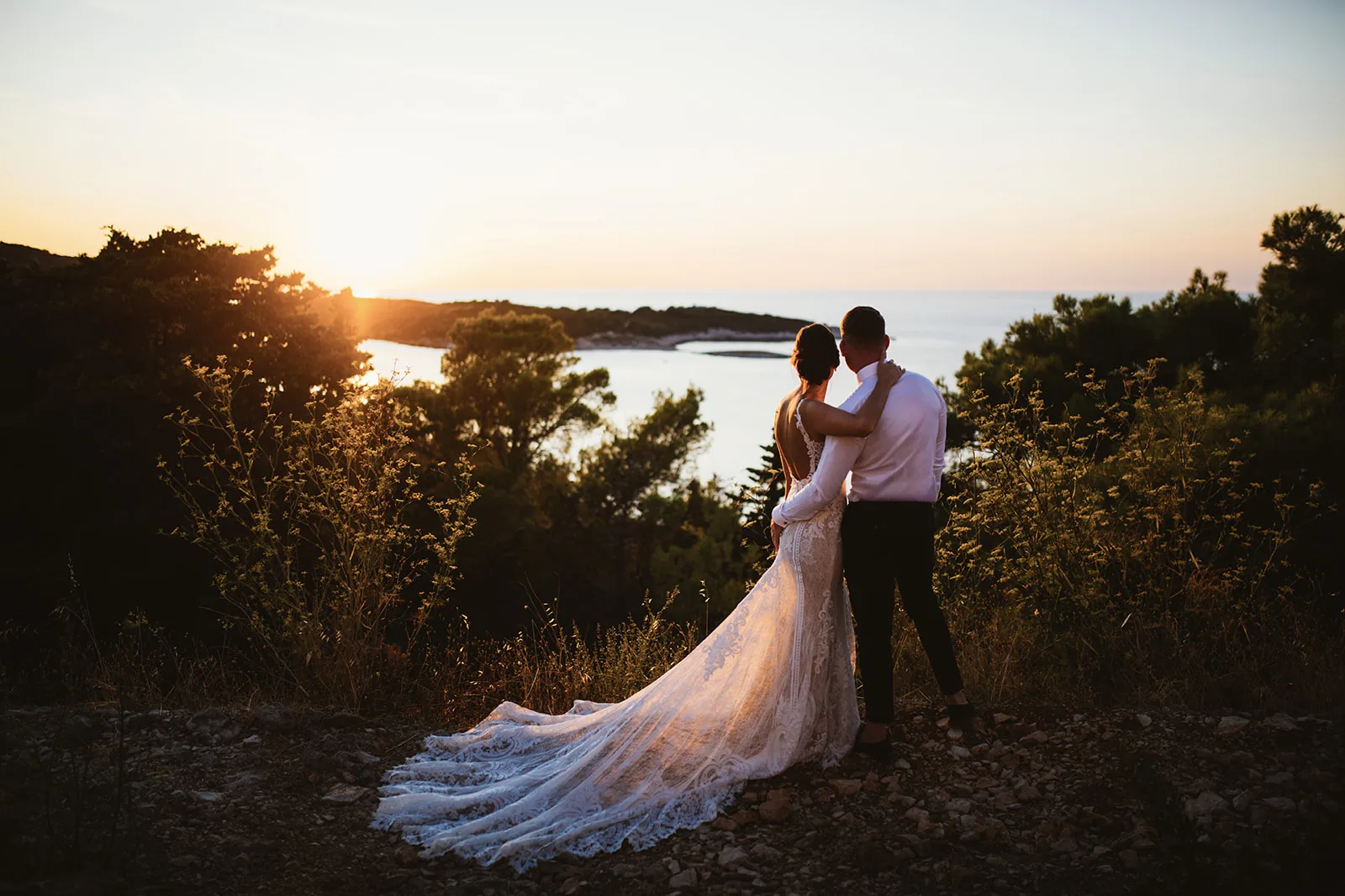 The couple stands wrapped together on a wild, scrub-covered clifftop, gazing out over a golden sunset bay dotted with small islands.