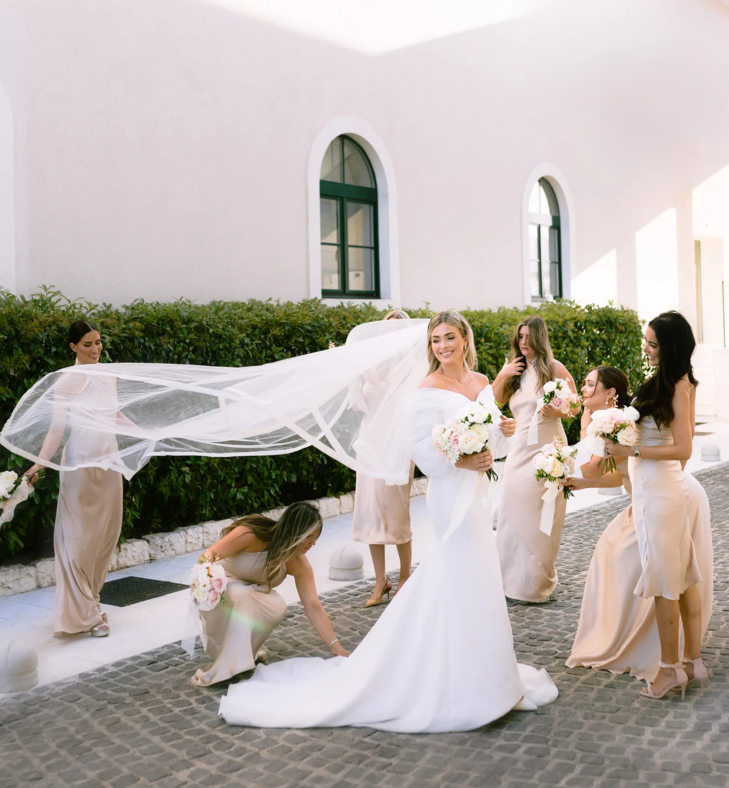 The bride walks forward smiling in an off-shoulder gown as her long veil billows sideways, held aloft by one bridesmaid while another kneels to arrange her train; three more bridesmaids in matching champagne satin flank her, each holding a bouquet.