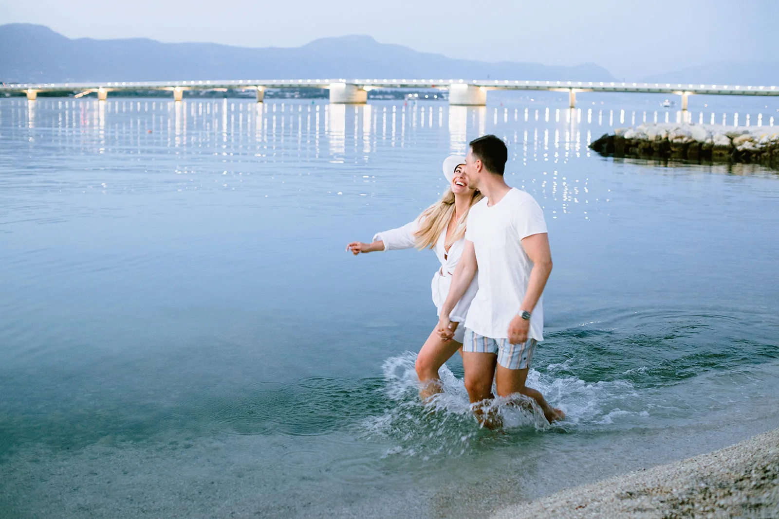 A couple splashes hand in hand through shallow water at dusk, a lit bridge glowing across the bay behind them, playful and relaxed.