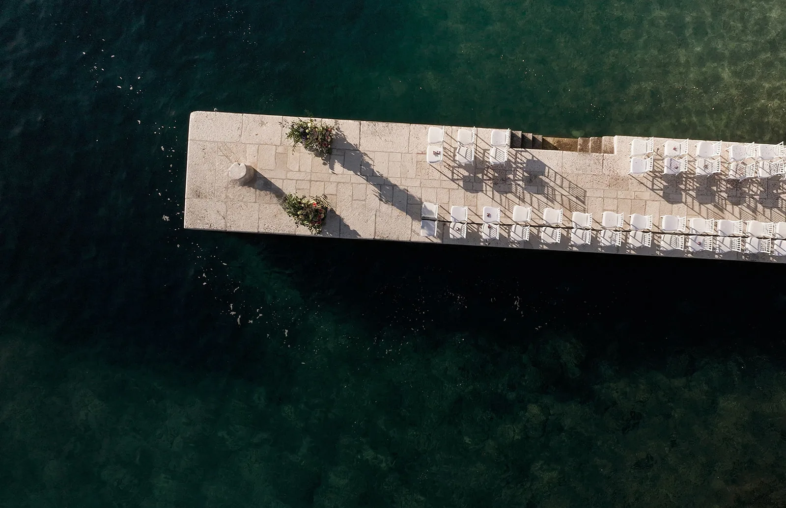 Straight-down drone view of a stone jetty extending diagonally into deep teal sea, set with two rows of white ceremony chairs and a pair of floral arrangements marking the aisle's end.