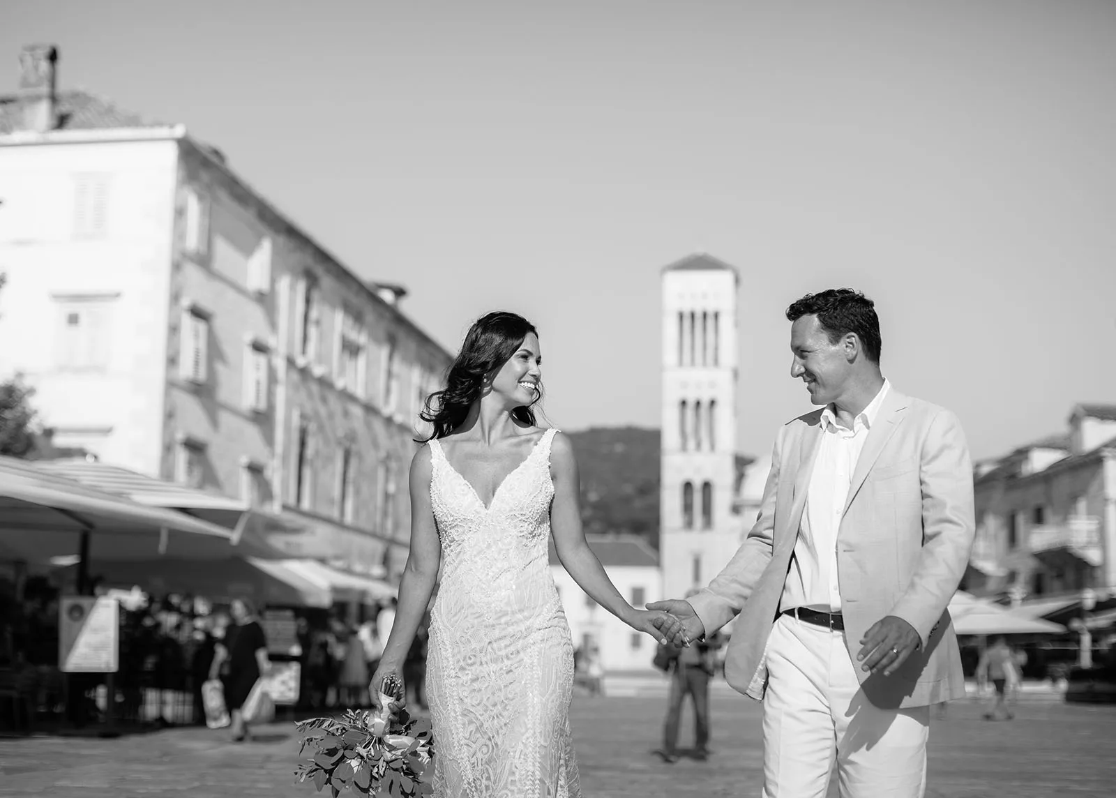 The bride and groom walk hand in hand across Hvar's main square, smiling at each other, her lace gown catching the breeze; the town's stone bell tower rises behind them among market umbrellas and passersby.