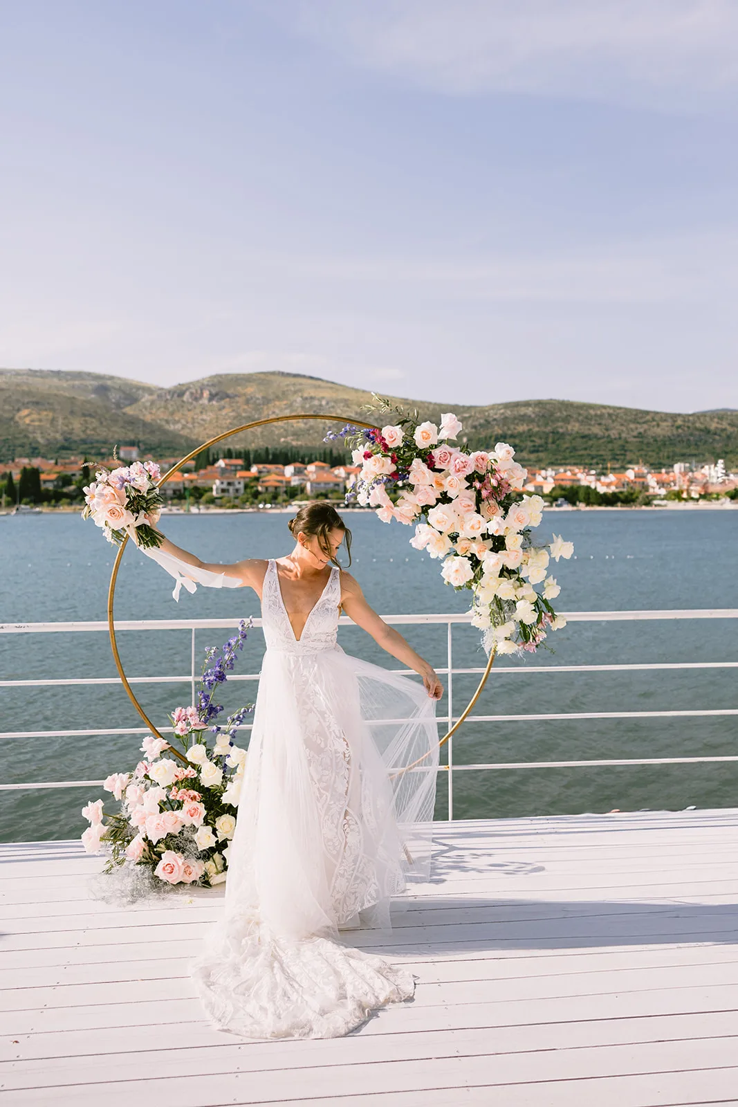 The bride poses inside a flower-wrapped gold hoop on a white dock, her gown catching the breeze, with a coastal town of red rooftops spread across the water behind her.
