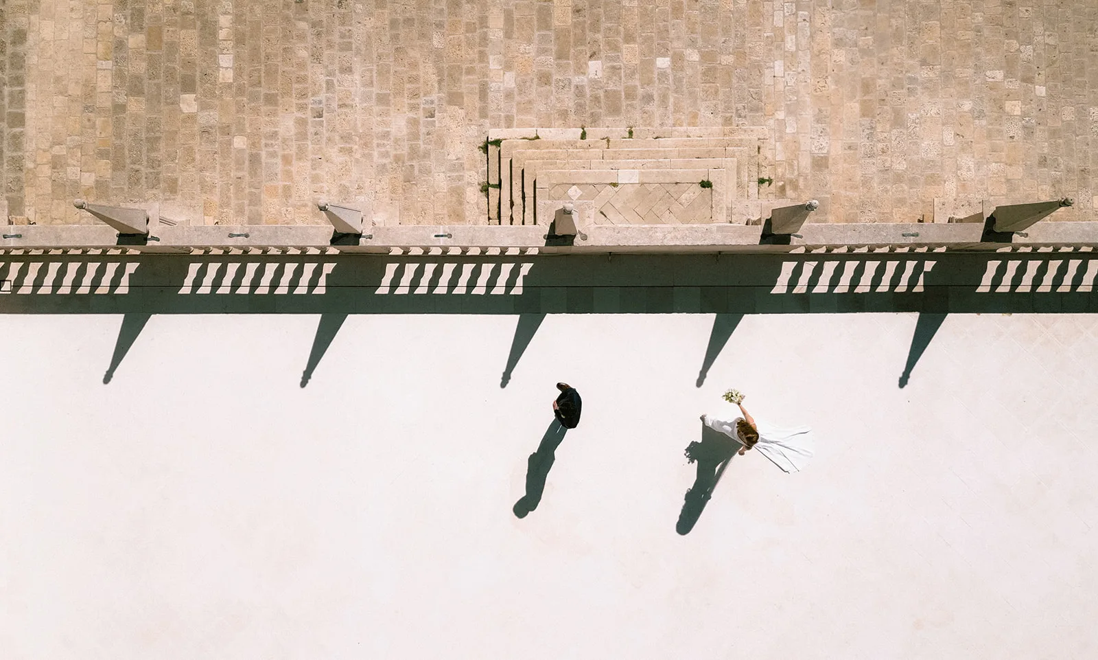 Directly overhead, the bride and groom walk along a sun-bleached stone terrace lined with cannon-mounted balustrades, their long shadows stretching across the pavement; stone steps rise toward the fortress wall behind them.