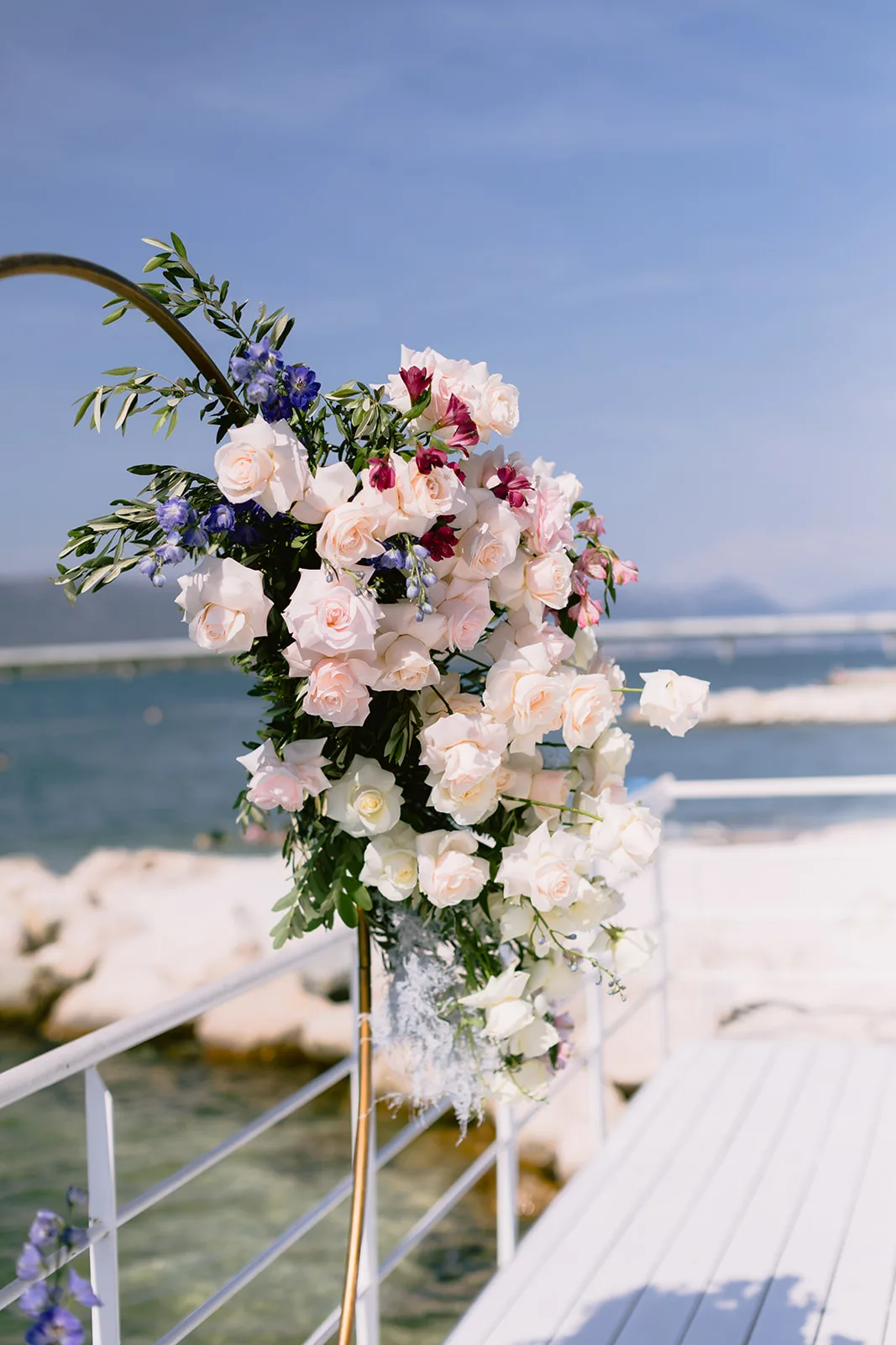 A cascading arrangement of blush roses and delphinium decorates a white dockside railing under a bright blue midday sky.