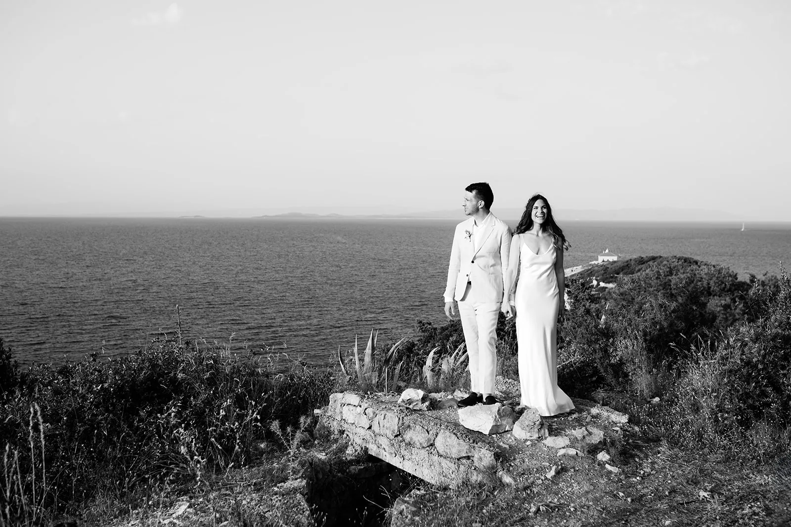 The bride and groom stand hand-in-hand on a low stone ruin atop a coastal hill, looking out over the sea; a small white building sits on a distant headland and a sailboat crosses the horizon.