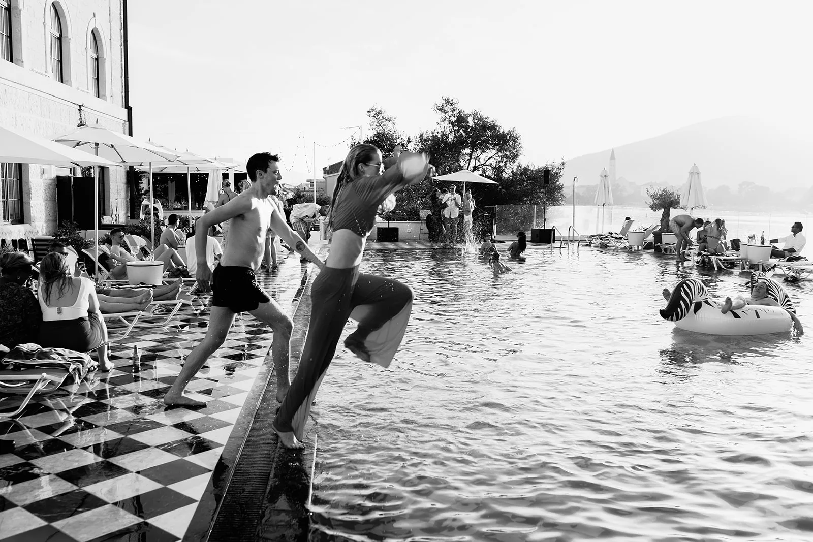 Two guests leap hand in hand off the pool's edge in a burst of motion, sunlit spray and a lively poolside crowd behind them, black and white.
