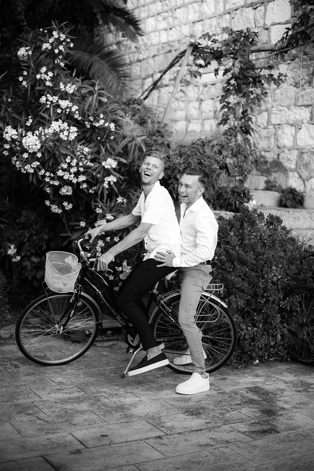 In black and white, two grooms ride off laughing on a bicycle through a flowering garden alley beside an old stone wall, caught in soft open shade.