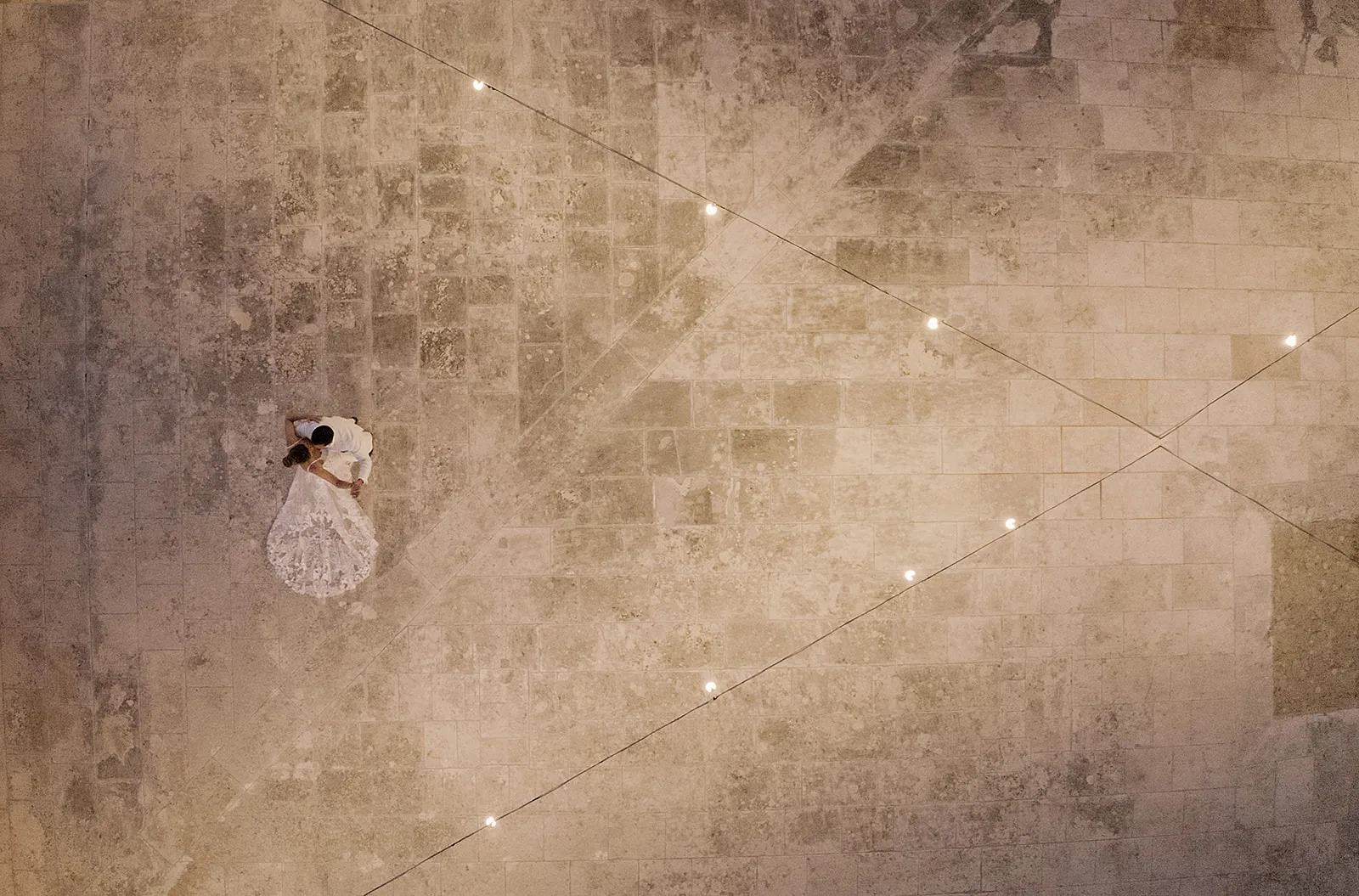 Directly overhead at dusk, the bride and groom embrace in the corner of a vast, worn limestone square; string lights cross the frame in sharp diagonal lines, dwarfing the couple in the open stone expanse.