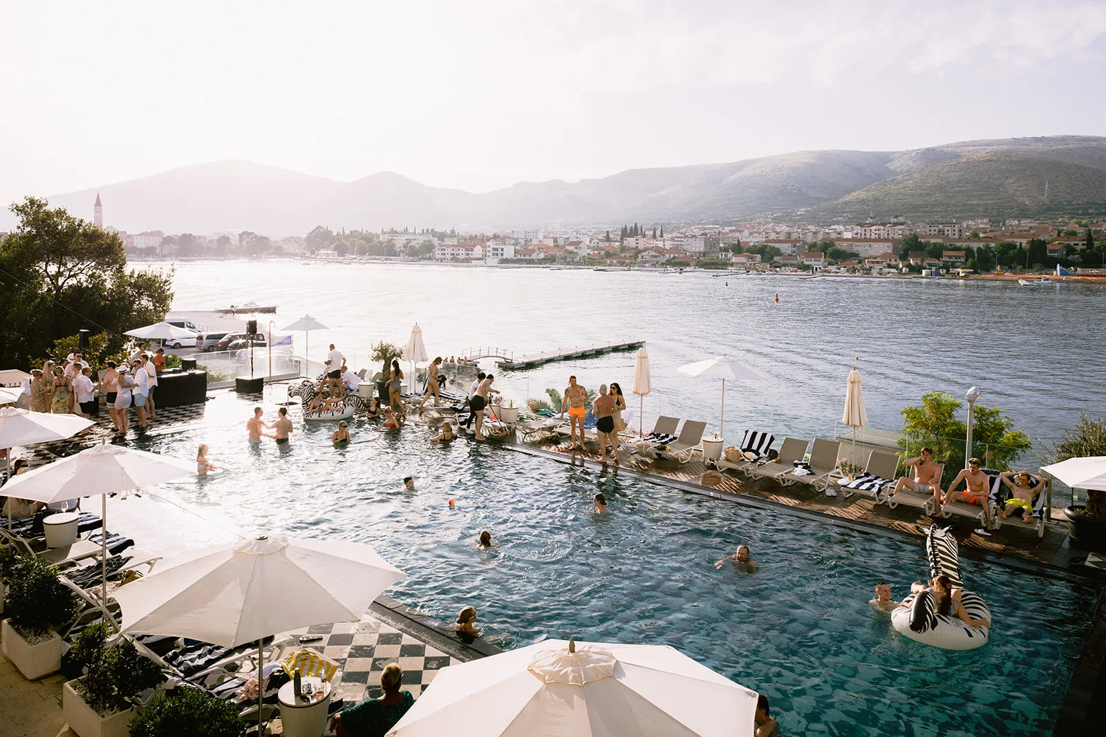 Wedding guests fill the infinity pool and sun deck of a coastal resort, floating on zebra-striped inflatables with a hillside town spread across the bay.