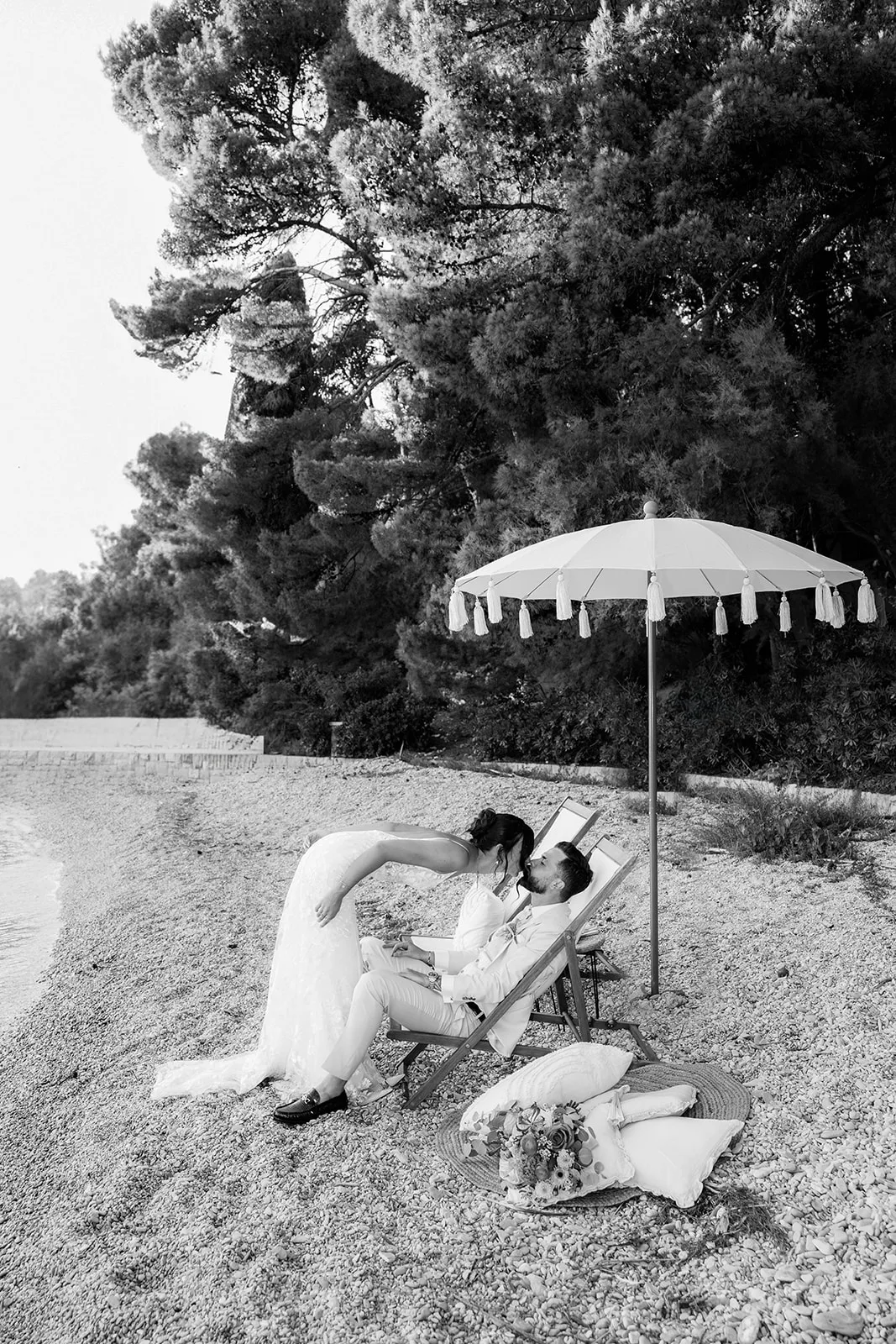 Black and white beach scene at dusk: the bride leans down to kiss the groom as they relax in deck chairs under a tasseled umbrella, pillows scattered on the pebbles.