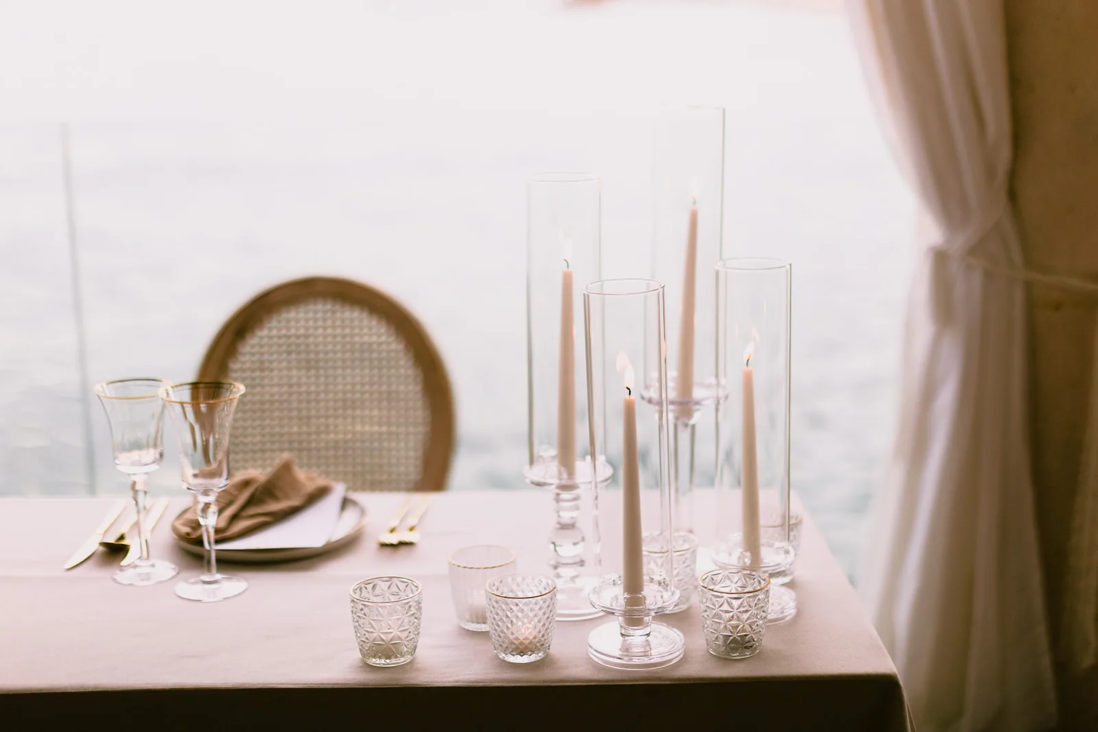 A cluster of lit taper candles in graduated clear glass hurricanes and faceted votives is arranged on a linen-topped table beside a sheer curtain, with gold-rim wine glasses and a folded napkin at the place setting behind.