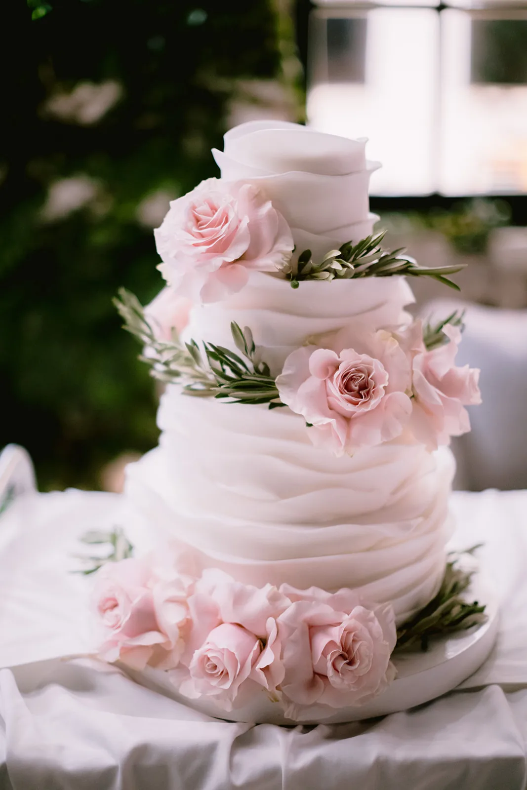 A ruffled white fondant wedding cake trimmed with blush garden roses and greenery stands ready on the dessert table, soft window light.