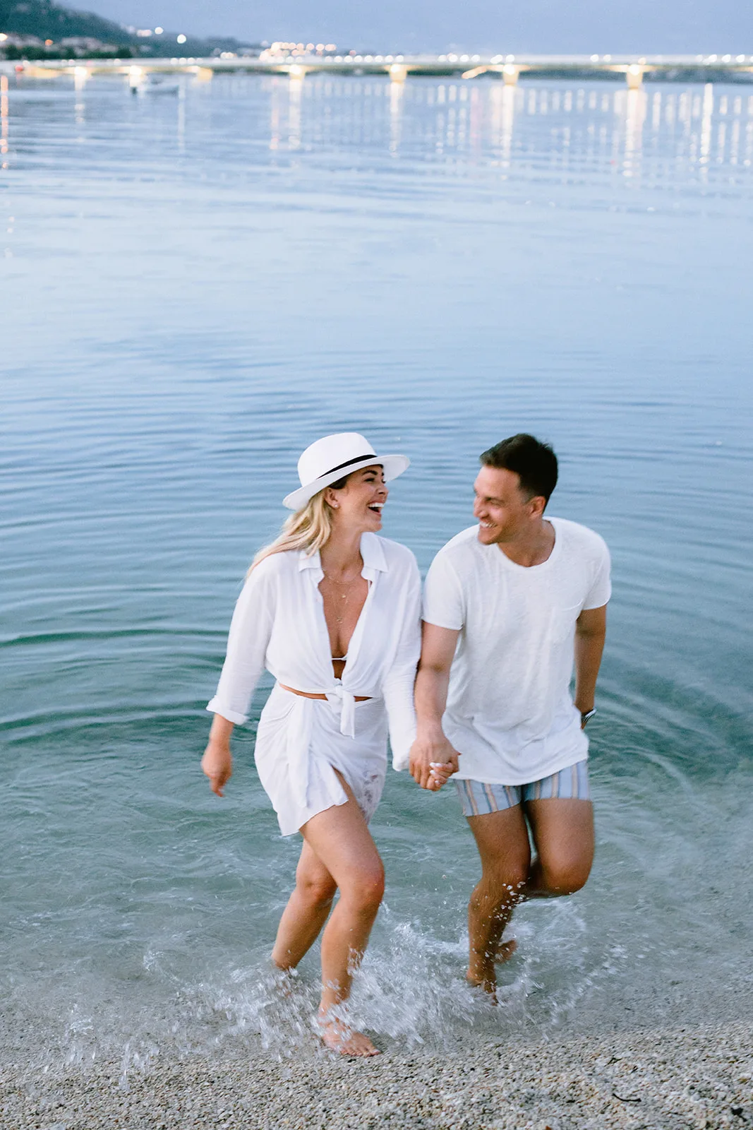 A couple laughs and holds hands while wading through shallow water at dusk, splashing as they walk; a brightly lit bridge and waterfront glow on the horizon behind them.