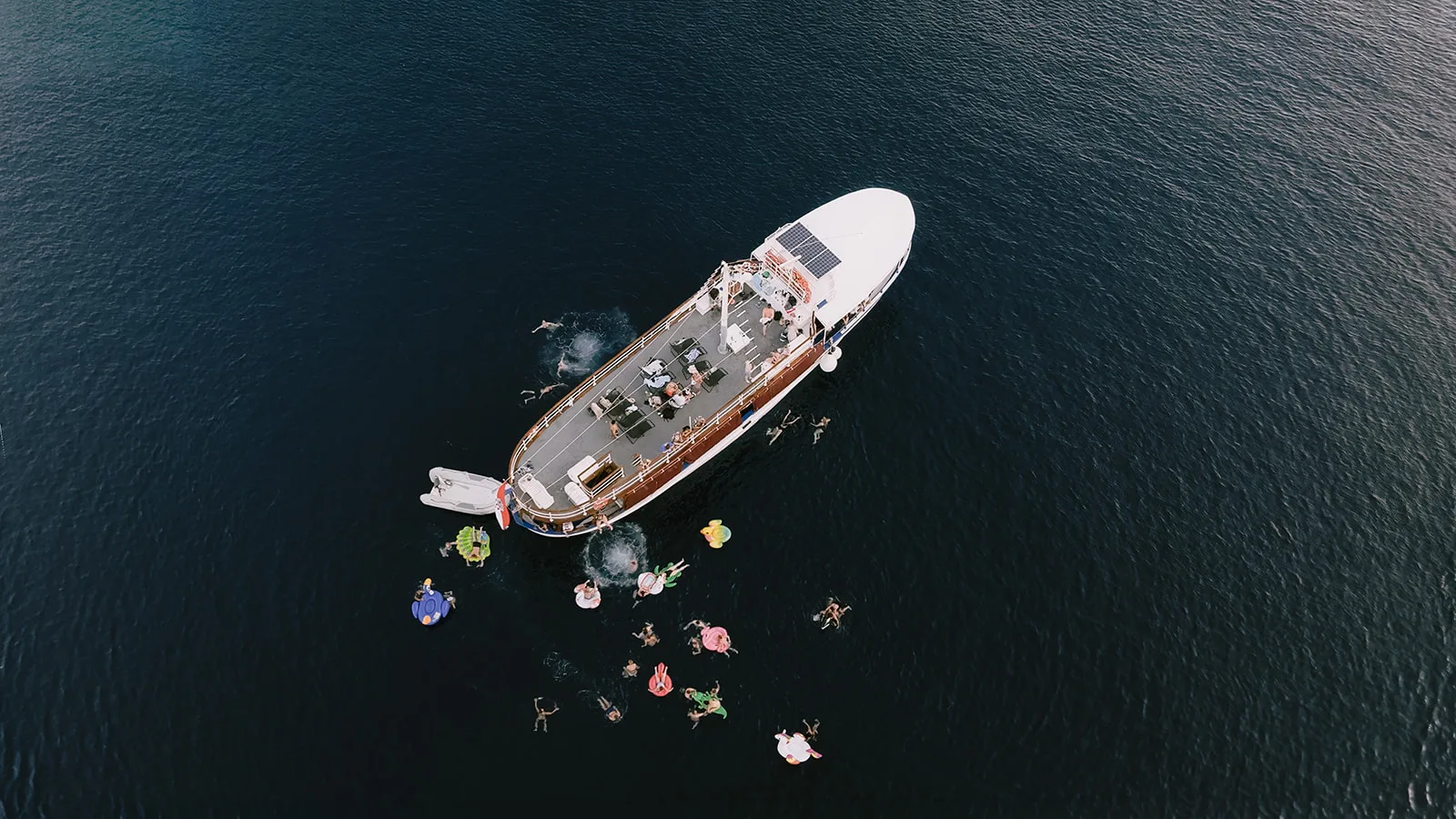 A drone shot looks straight down on a wooden charter boat anchored in deep blue water, guests sunbathing on deck and swimming among colorful inflatables.