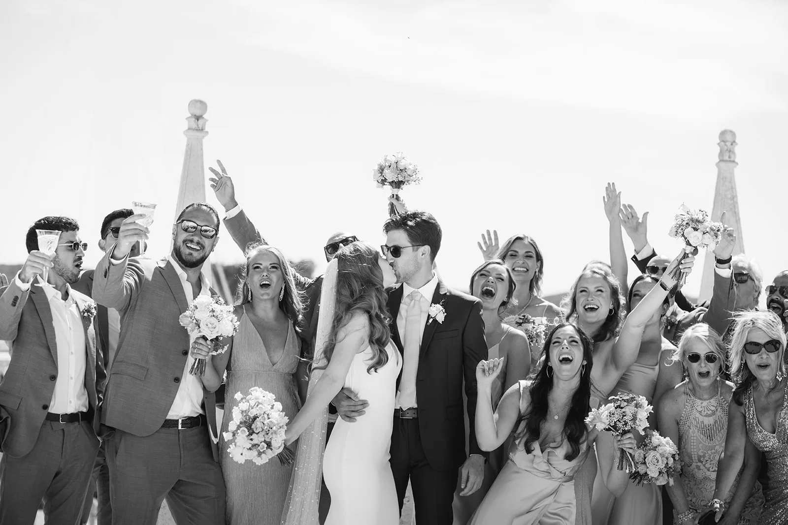 Black and white: the bride and groom kiss while their wedding party erupts around them, raising champagne and cheering under a bright open sky by the water.