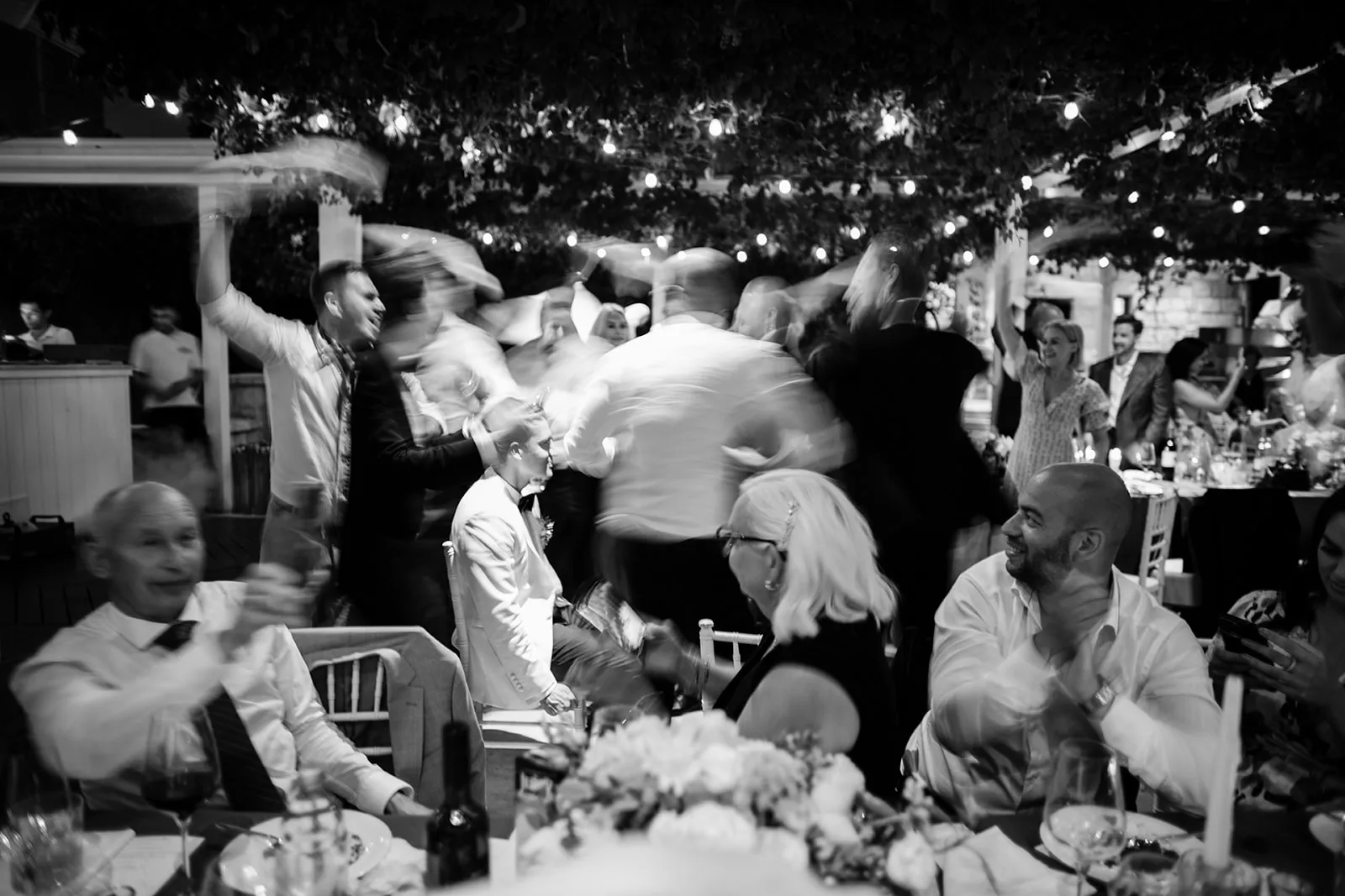 Black and white: a blur of guests dance under string-lit trees while, at the table in the foreground, others laugh and clap along to the music.