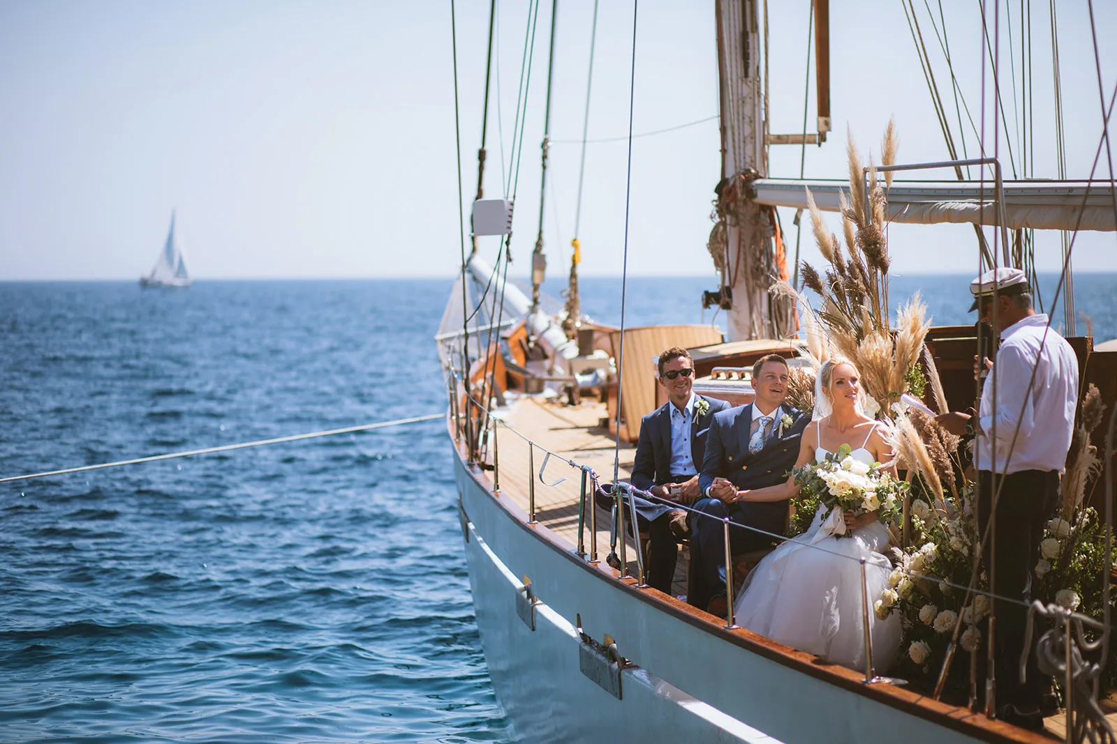The bride and groom sit hand-in-hand at the bow of a wooden sailboat with a groomsman beside them, as an officiant stands at the rail; pampas grass and white roses decorate the bow railing.