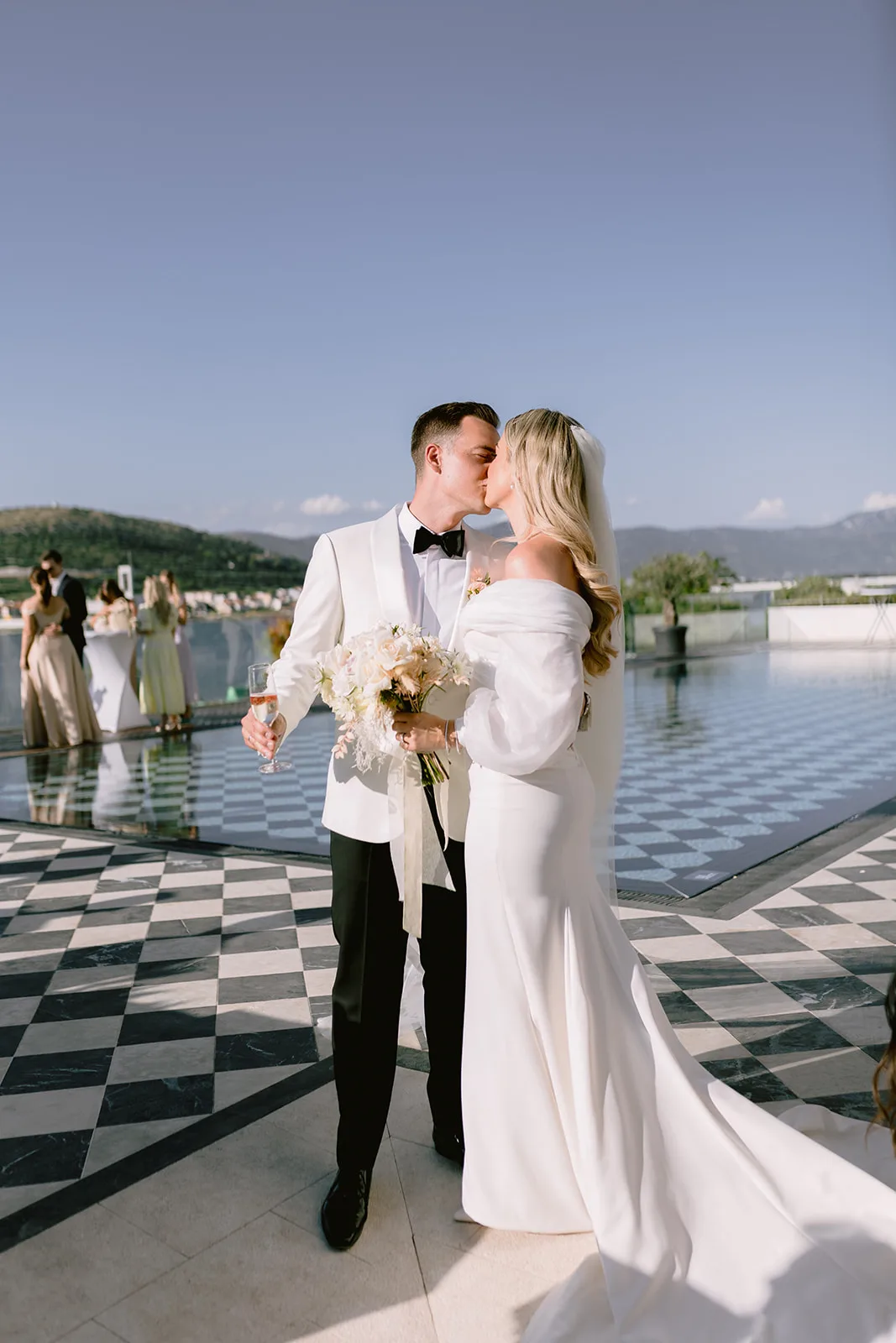 Bride and groom kiss on a black-and-white checkerboard terrace beside an infinity pool, he in a white dinner jacket holding a champagne flute, she in an off-shoulder gown holding her bouquet.