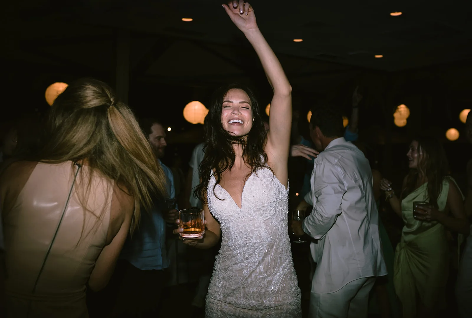 The bride throws her arm up and laughs mid-dance, drink in hand, surrounded by guests on a lantern-lit dance floor.