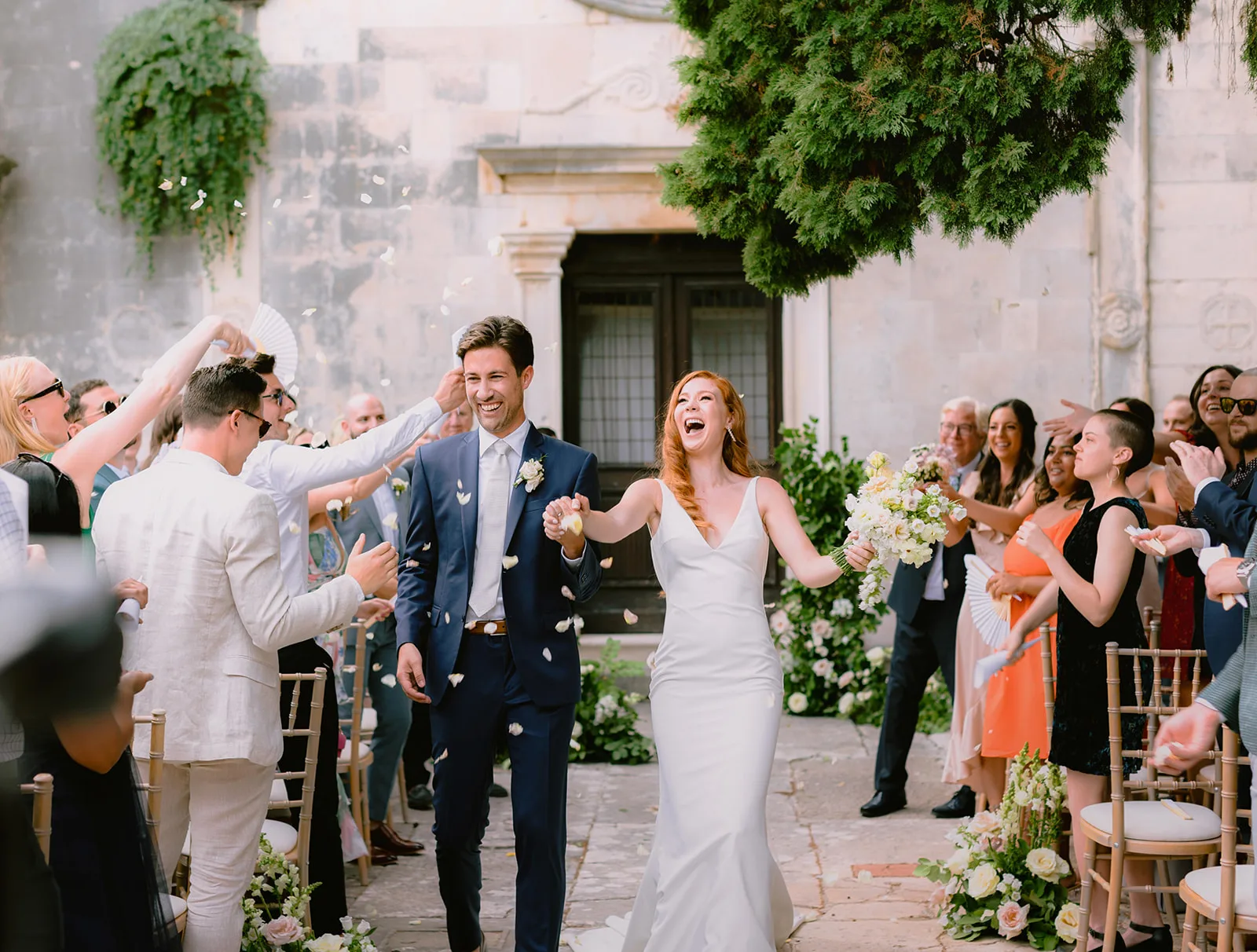 The newlyweds walk beaming through a shower of confetti as cheering guests line the stone church courtyard, bright midday sun.