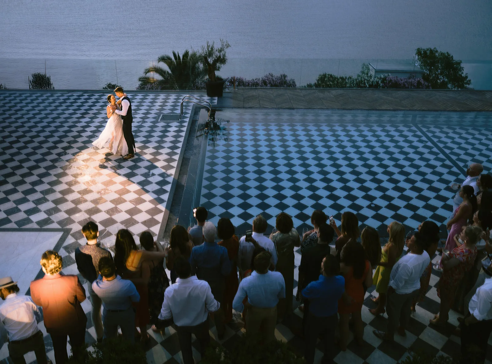 Shot from above, a bride and groom share their first dance in a warm spotlight on a checkered terrace, guests ringed around them at dusk.