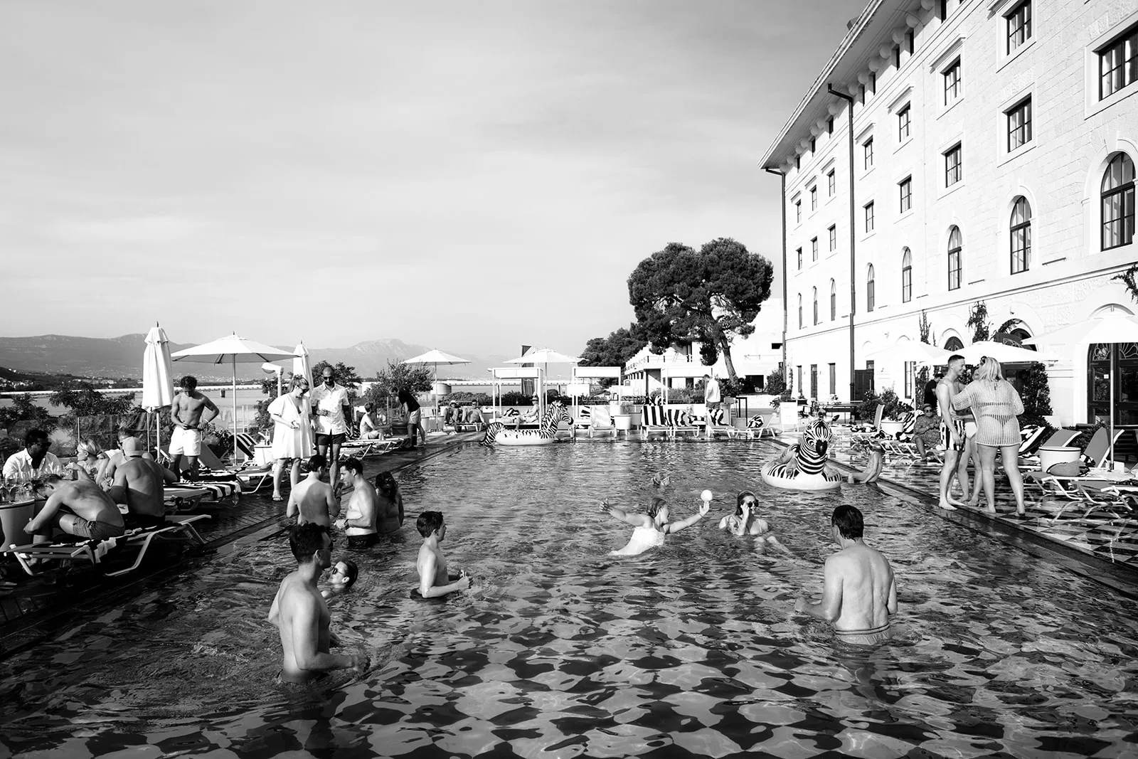 A crowd of guests splashes and plays in a large hotel pool, the bride visible among them still in her white dress, as zebra-print inflatables float nearby and others lounge poolside under umbrellas.