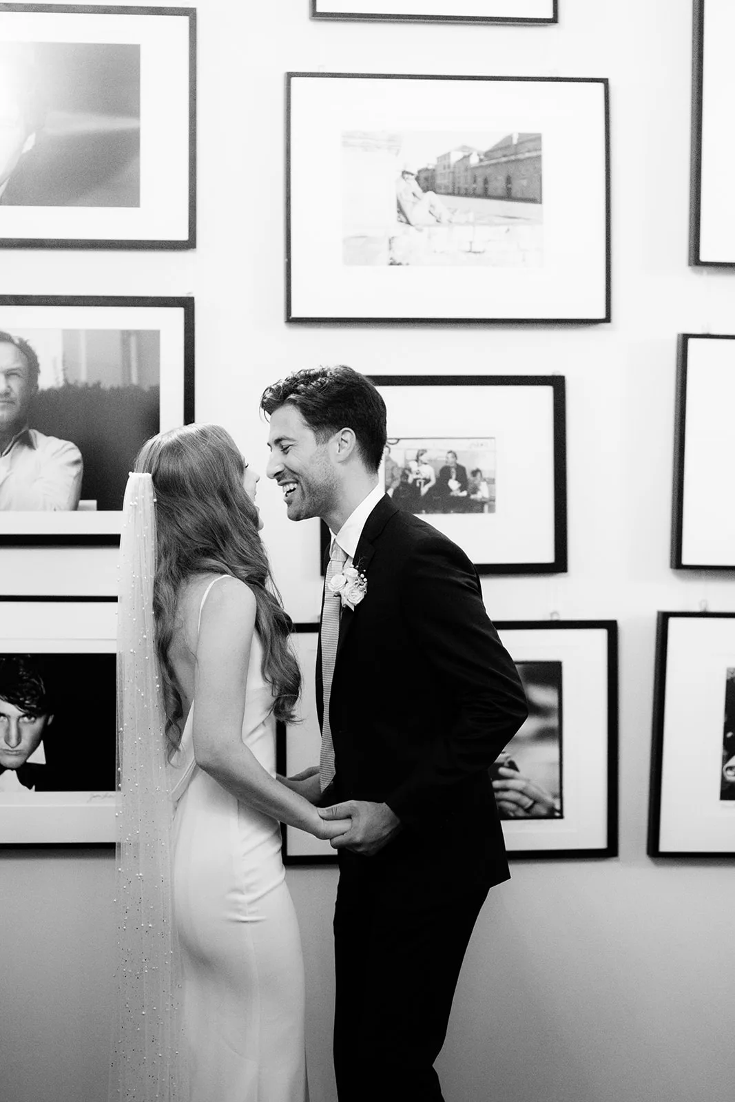 Black and white: the bride and groom laugh together, foreheads close and hands clasped, standing inside a gallery walled floor-to-ceiling with black-and-white photography, reminiscent of Dubrovnik's War Photo Limited.