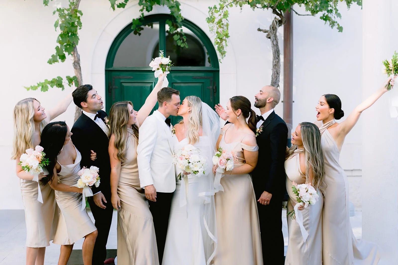 The bride and groom share a kiss at the center of their wedding party, bridesmaids in champagne satin and groomsmen in black tuxedos cheering around them, in front of a white building with an arched green door and grapevine overhead.
