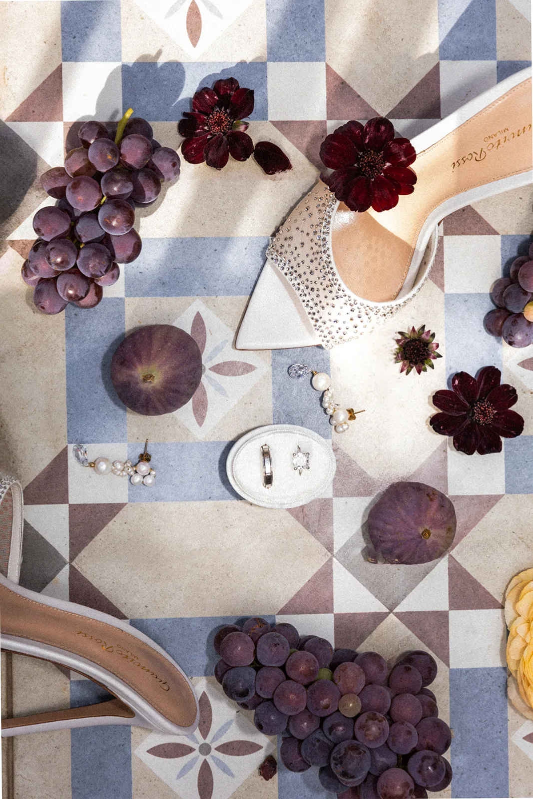 A sun-drenched flat lay on patterned Mediterranean tile brings together crystal-studded heels, dark grapes and figs, dahlias and the wedding rings.
