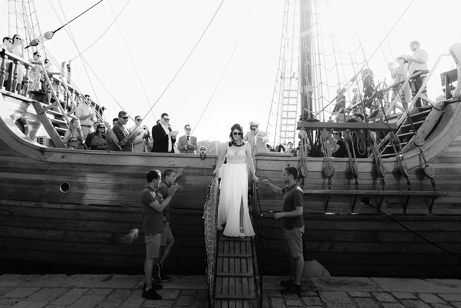 The bride walks down a wooden gangway between two tall wooden ships moored in a historic harbor, crew members steadying the ropes on either side while guests aboard both vessels applaud.