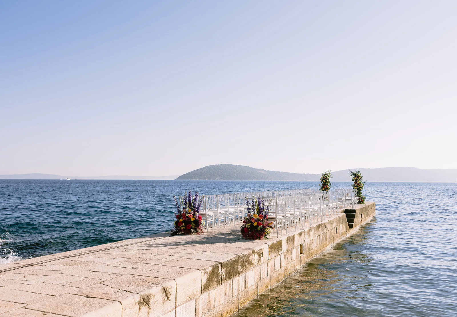 A ceremony aisle of white chairs stretches down a narrow stone pier straight into the sea, framed by two colorful floral arches with island silhouettes on the horizon.