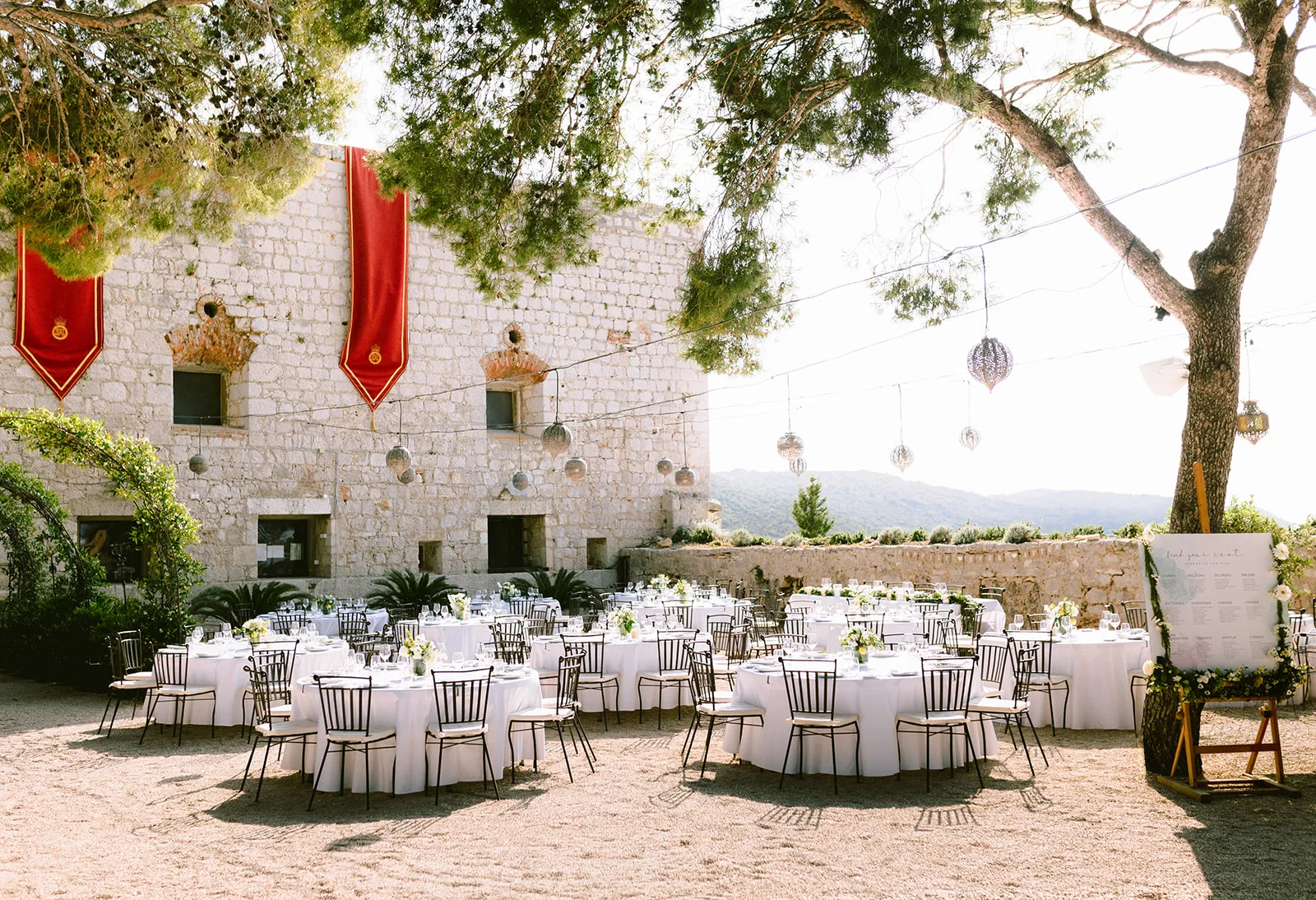 An empty reception courtyard, round tables dressed in white linen beneath a canopy of pine and hanging lanterns, red heraldic banners draped against pale stone walls, hills rolling away in the soft afternoon haze.