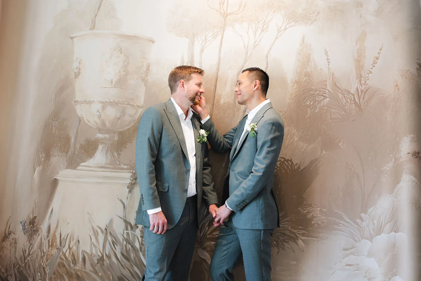 Two grooms share a tender moment, one cradling the other's face, in front of a soft painted mural backdrop.