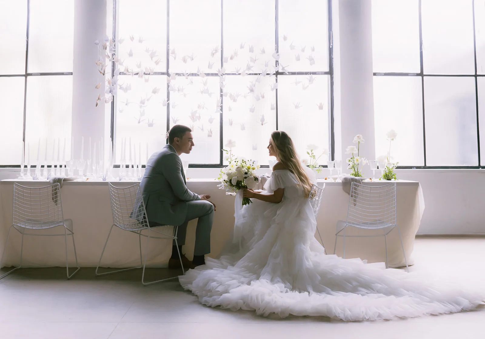 In a bright, airy industrial loft strung with paper cranes, the bride kneels in her tulle gown while the groom sits at the table's edge, facing each other quietly.