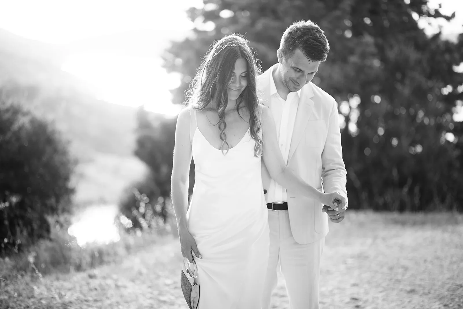 A black-and-white portrait of the couple walking close together along a gravel path, hazy backlight flaring behind them for a soft, romantic glow.