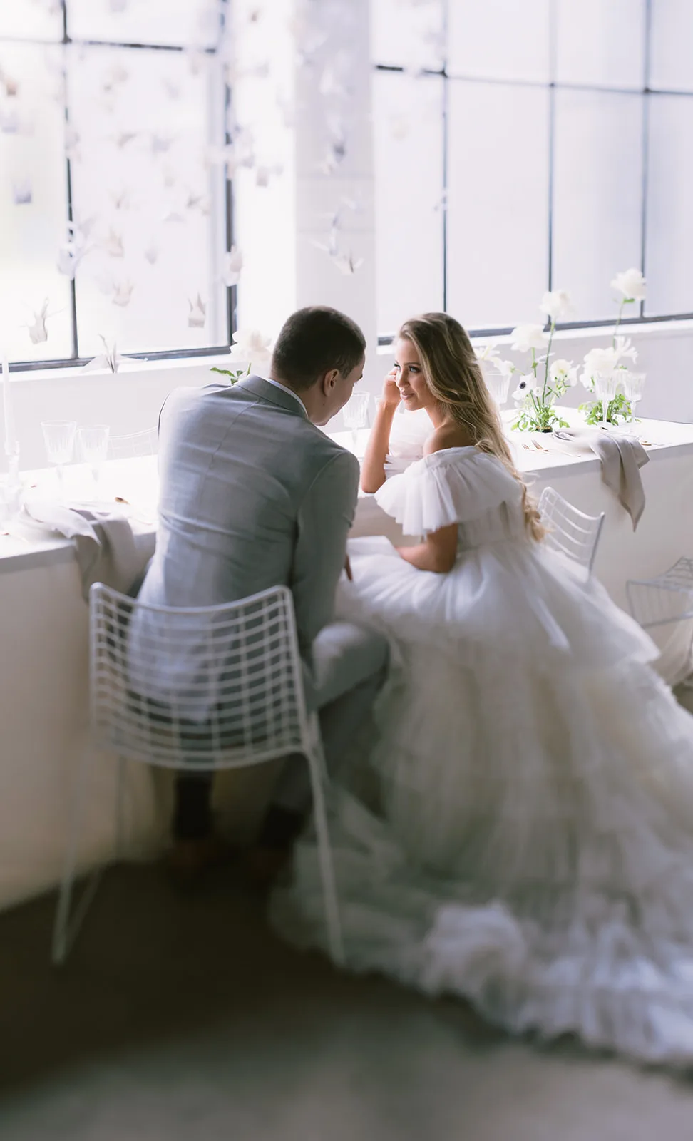 Seen from behind, the groom in a grey suit leans toward the bride as she touches her cheek and smiles, both seated at a narrow ledge table before a tall window strung with paper butterflies and floral sprigs.