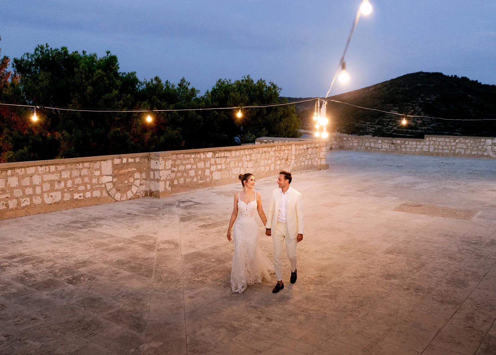 The newlyweds stroll hand in hand across a wide stone terrace at blue hour, string lights glowing overhead and terraced hills rolling behind them.