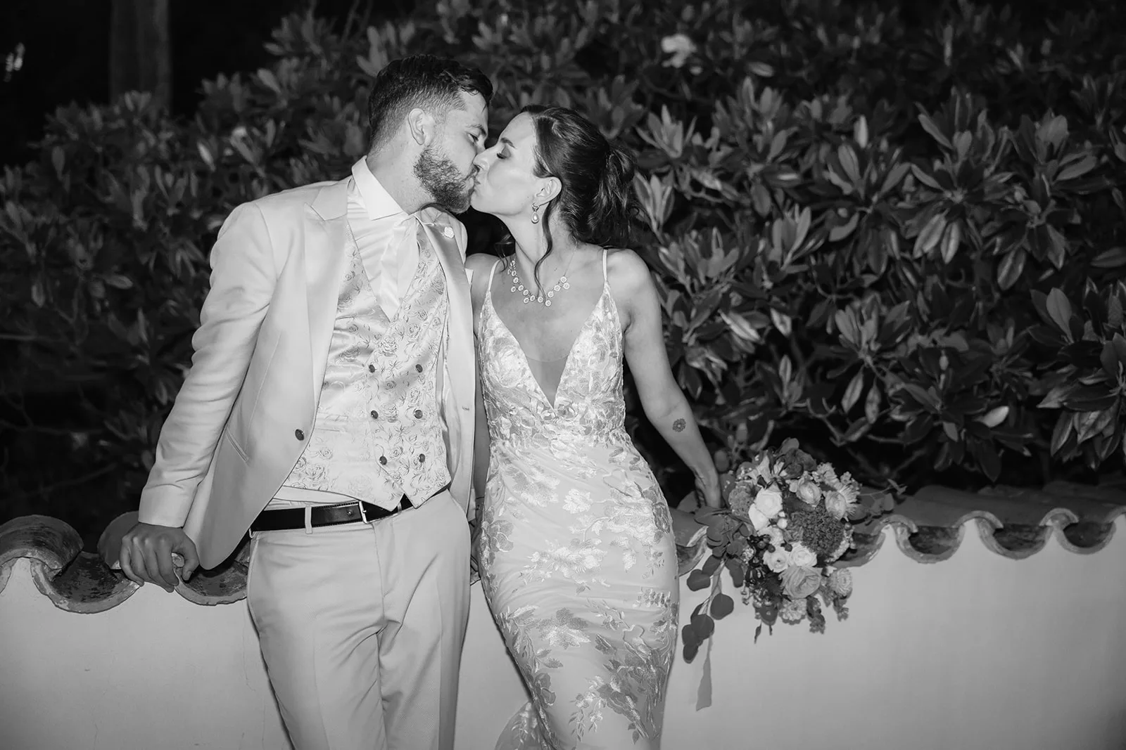 A black-and-white night portrait of the couple kissing against an ornate balustrade, her metallic floral gown and jewelry catching the flash, bouquet in hand.