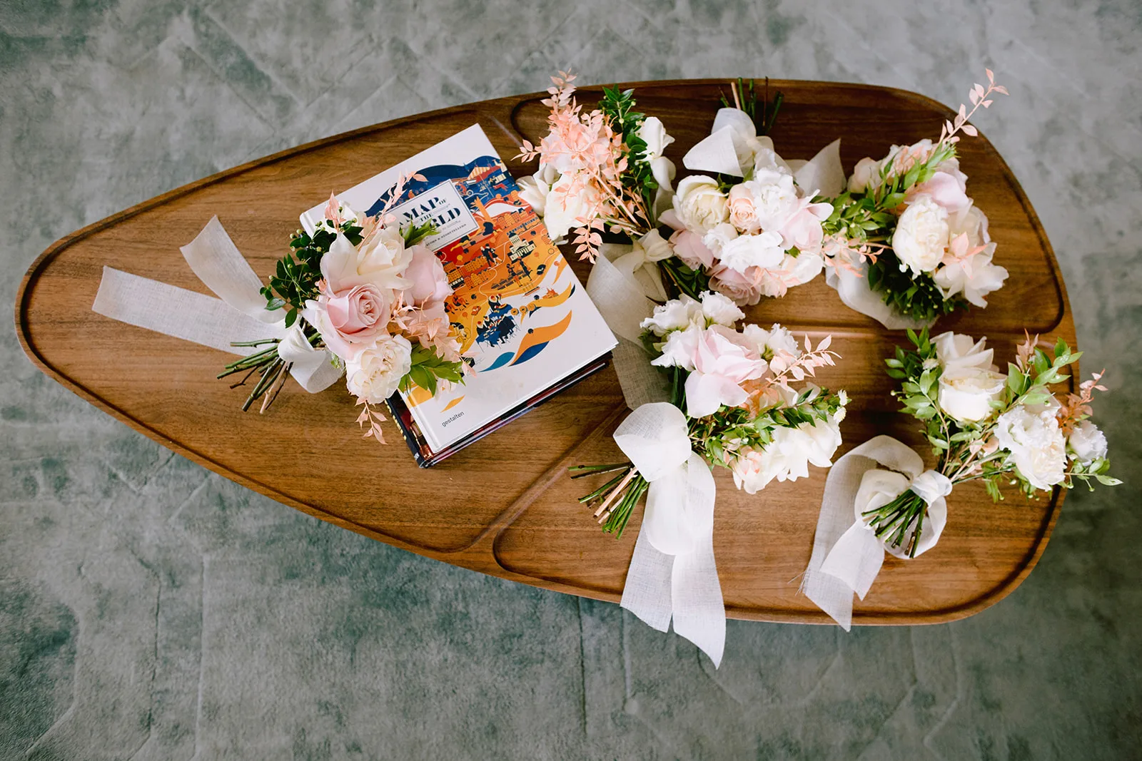 Five bridesmaid bouquets of blush and white roses, peonies, and eucalyptus, tied with trailing ivory ribbon, are arranged in a circle on a wooden surfboard-shaped coffee table beside an illustrated atlas book.