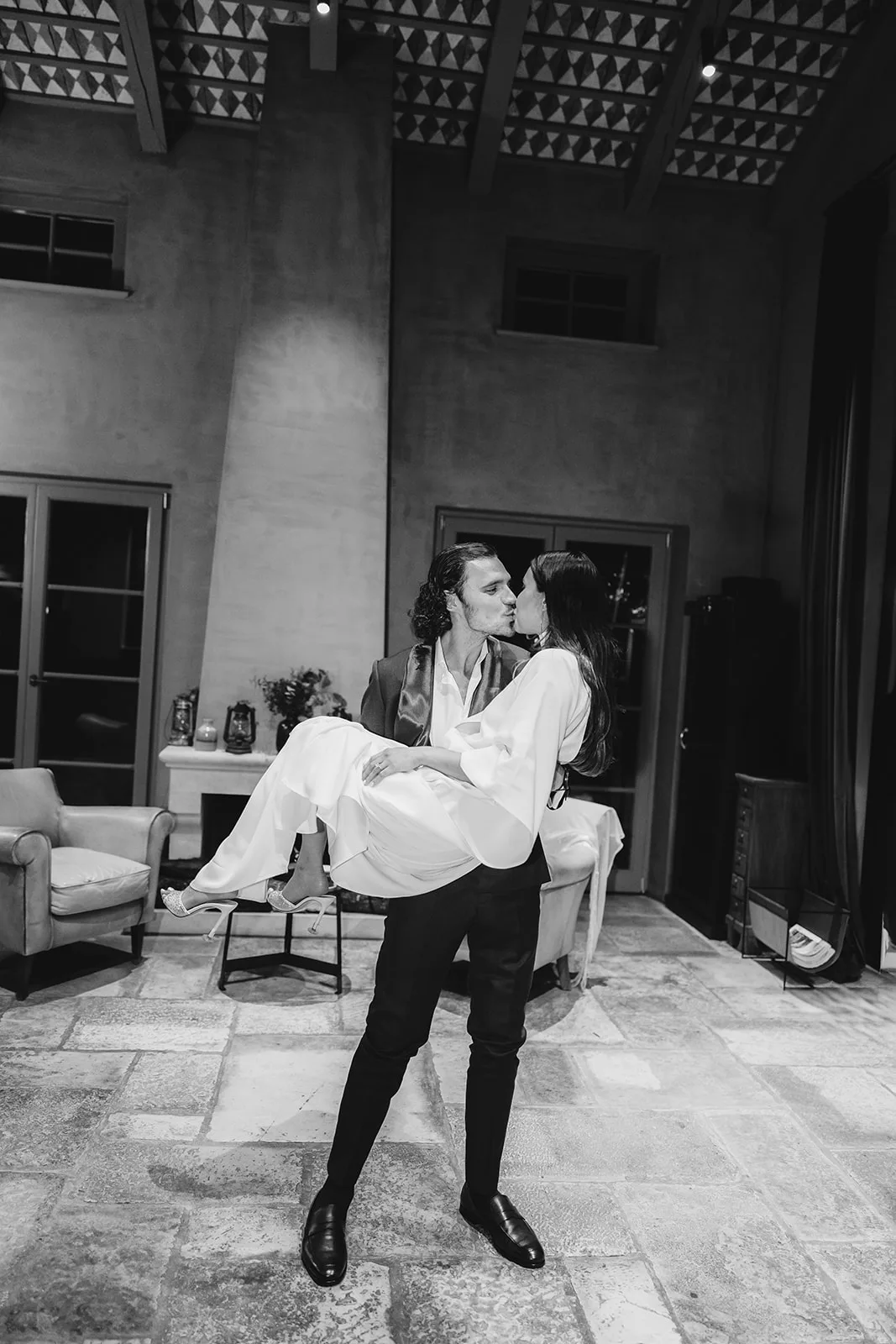 Black and white: the groom lifts and carries the bride, both kissing, through a vaulted stone-and-timber great room lit by warm lamplight and a quiet fireplace.