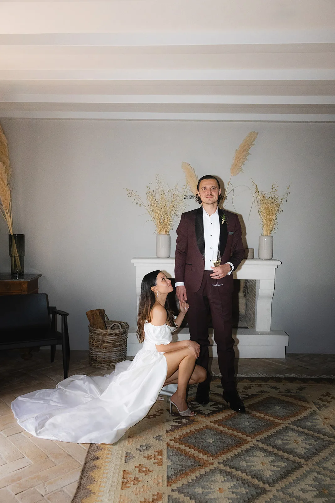 The bride kneels teasingly at her groom's feet as he stands by a stone fireplace with a glass of champagne, dried pampas grass and a kilim rug setting a relaxed, boho tone.