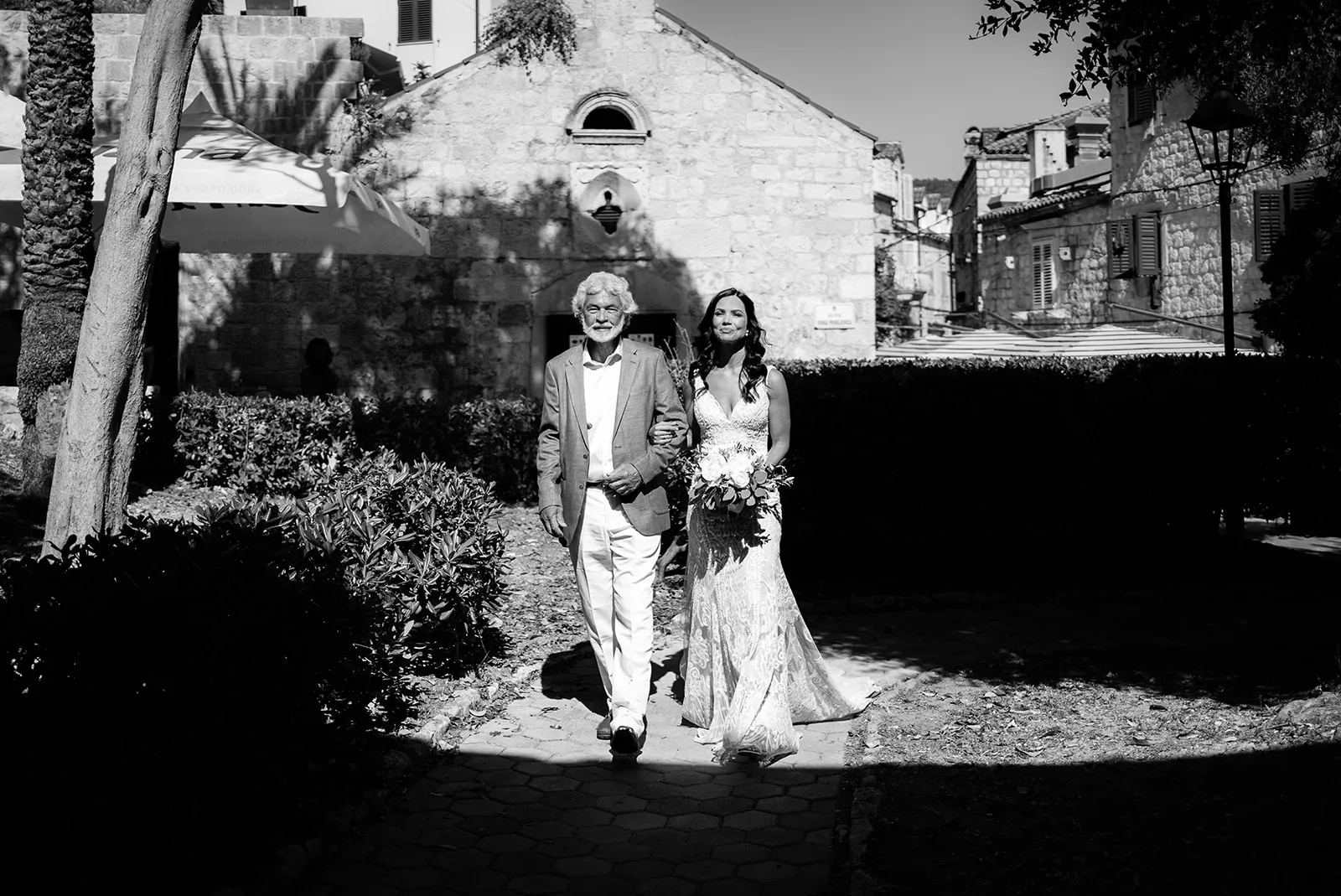 The bride's father walks her along a sun-drenched courtyard path toward a stone chapel, framed in a sharp pool of light amid deep shadow, black and white.