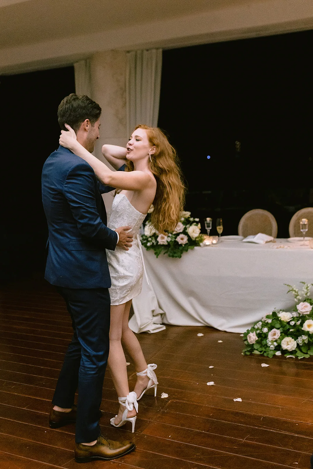 The newlyweds sway close on the dance floor at night, the bride laughing as she reaches up to the groom, their candlelit sweetheart table glowing behind them.