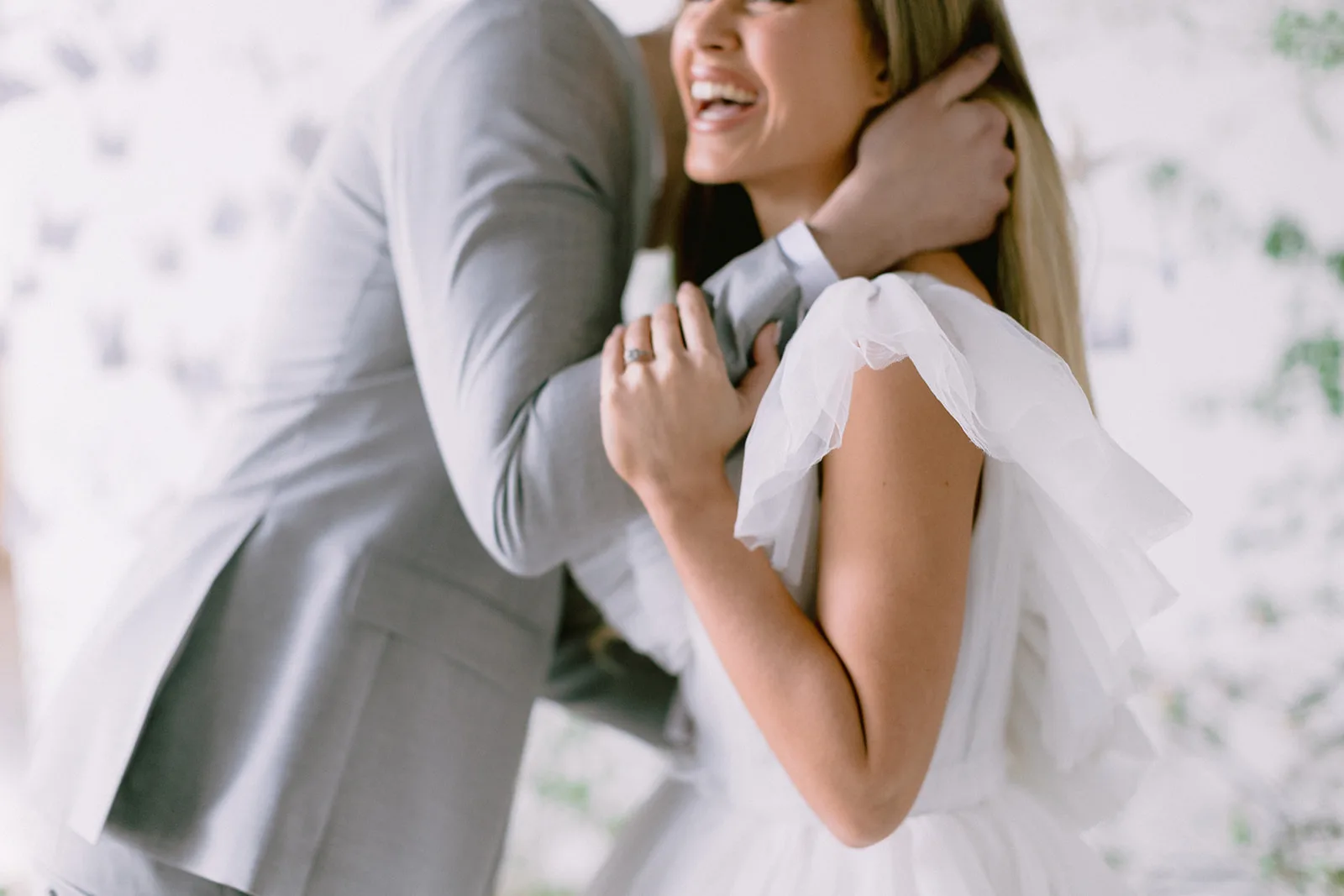 In soft, even interior light, the bride laughs as the groom wraps her in an embrace, her hand with its engagement ring resting against his lapel and a tulle bow catching on her shoulder.