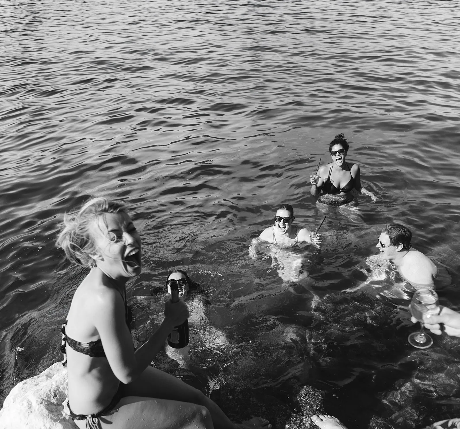 A guest in a bikini sits laughing on a limestone rock, opening a bottle toward the camera, while four more guests swim and laugh in the open sea around her holding cocktails and a raised champagne coupe.
